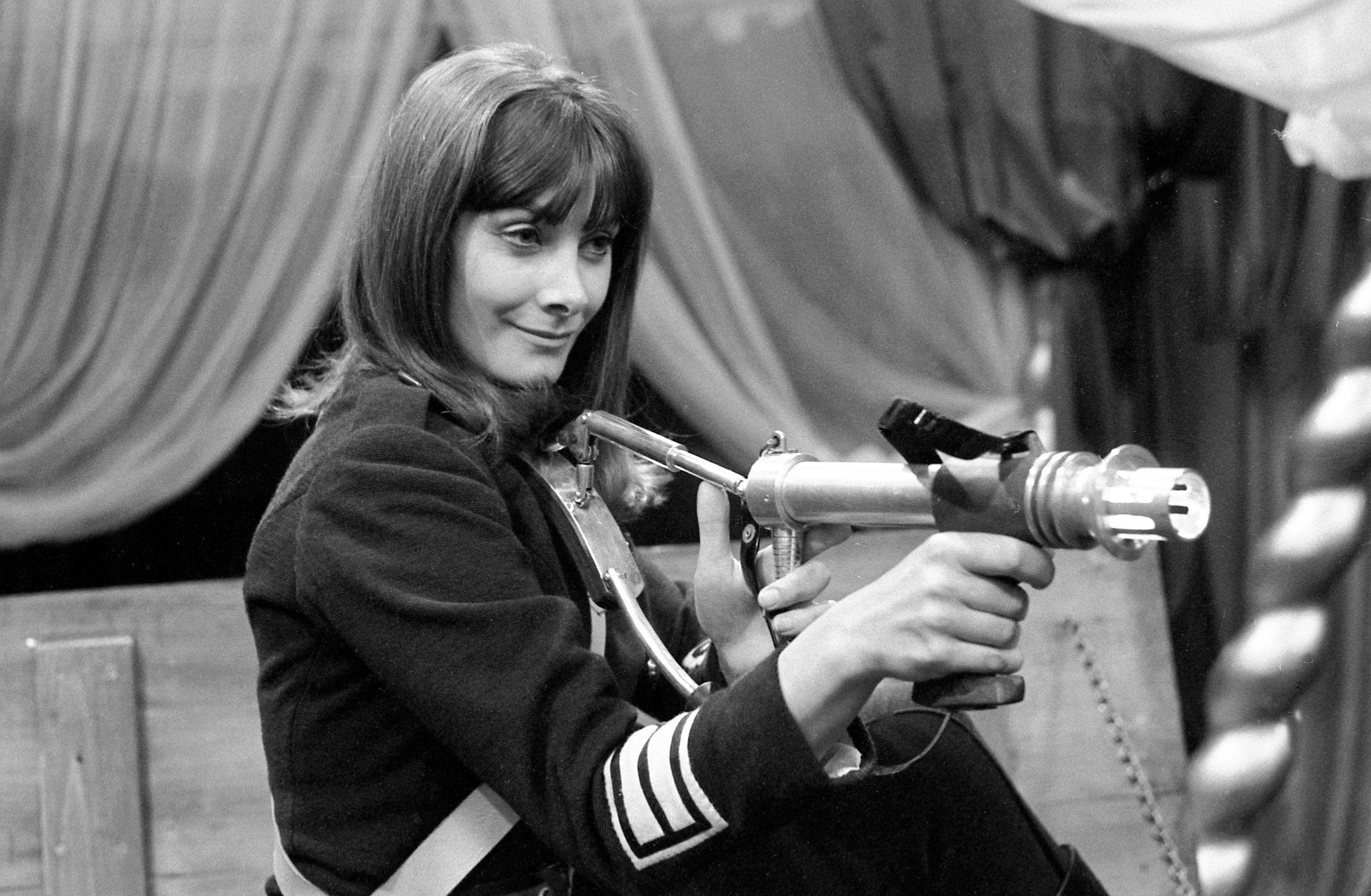 Black and white photo of Jean Marsh in character as Sara Kingdom on the set of The Daleks' Master Plan. She has long dark hair and wears a dark military style uniform with stripes on the right sleeve. She is holding a futuristic ray-gun weapon which is pointed o the right of the picture at someone out of shot. She has a smile on her face to suggest she is pleased with her actions.