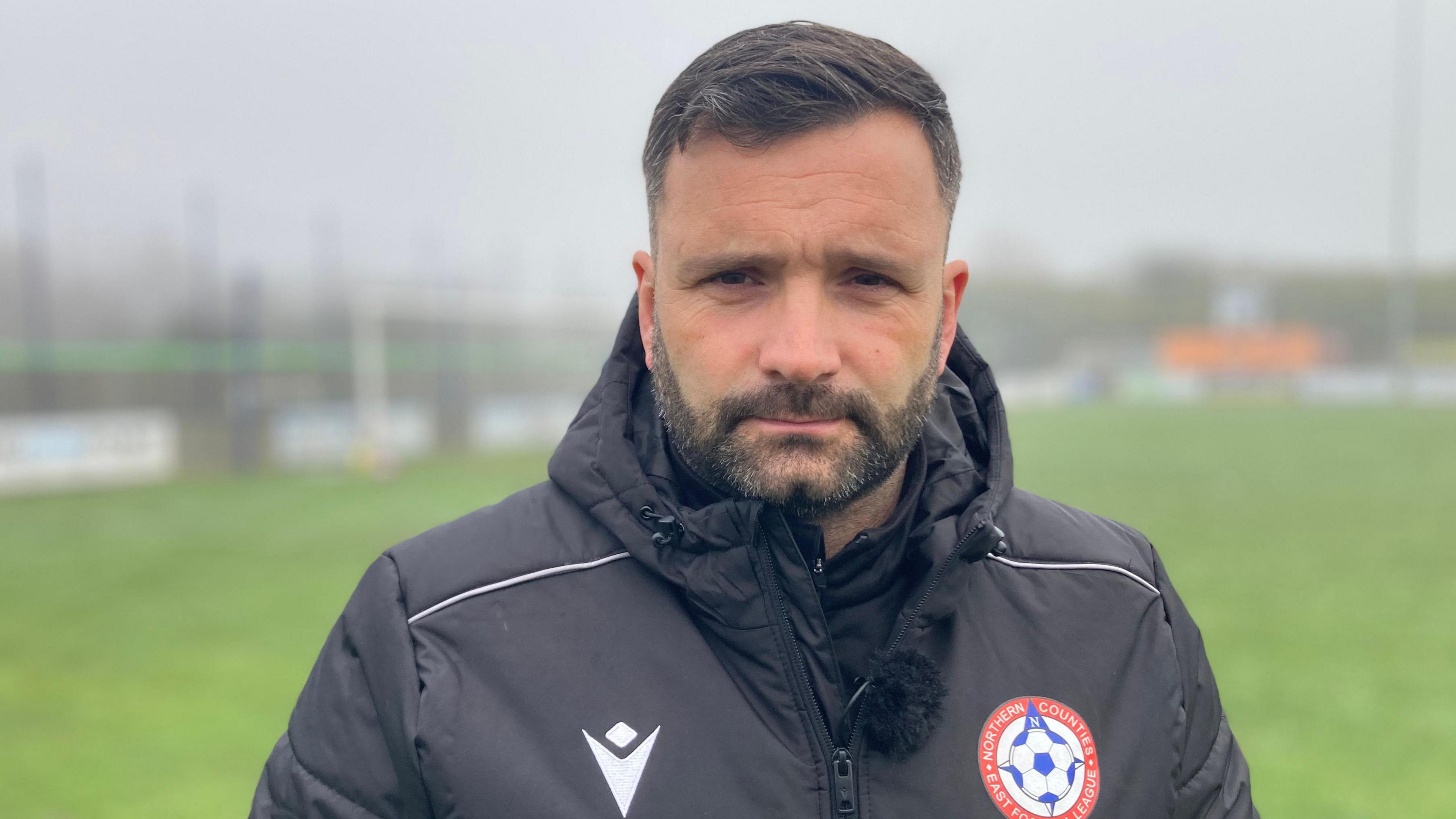 A picture of Anthony Bowsley standing on the pitch wearing a black jacket bearing the club's badge. Mr Bowsley has dark hair and a short beard.