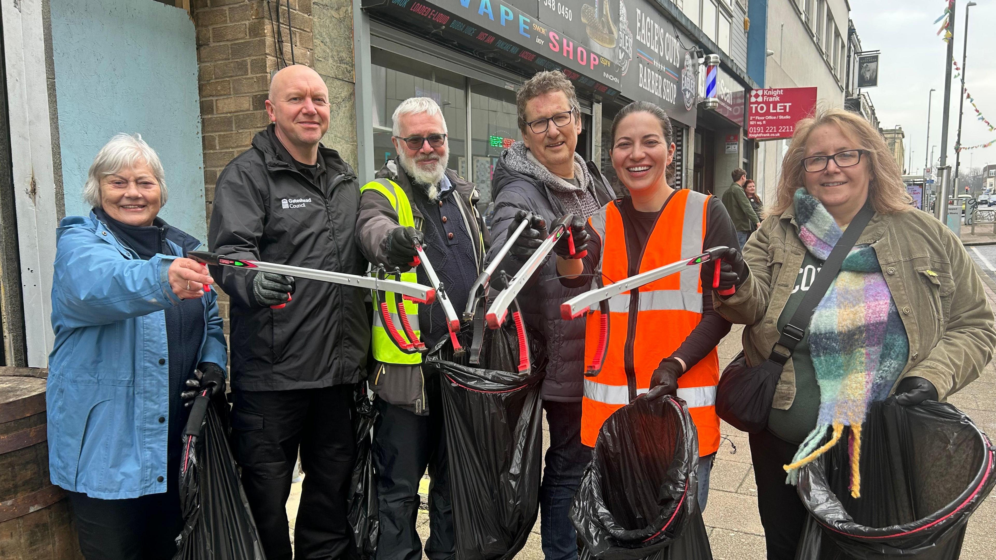 Luisa Scott among a group of five people with litter pickers and bin bags on Gateshead High Street. She has dark hair, tied back, and is wearing an orange hi-vis jacket. They are all looking directly into the camera and smiling.