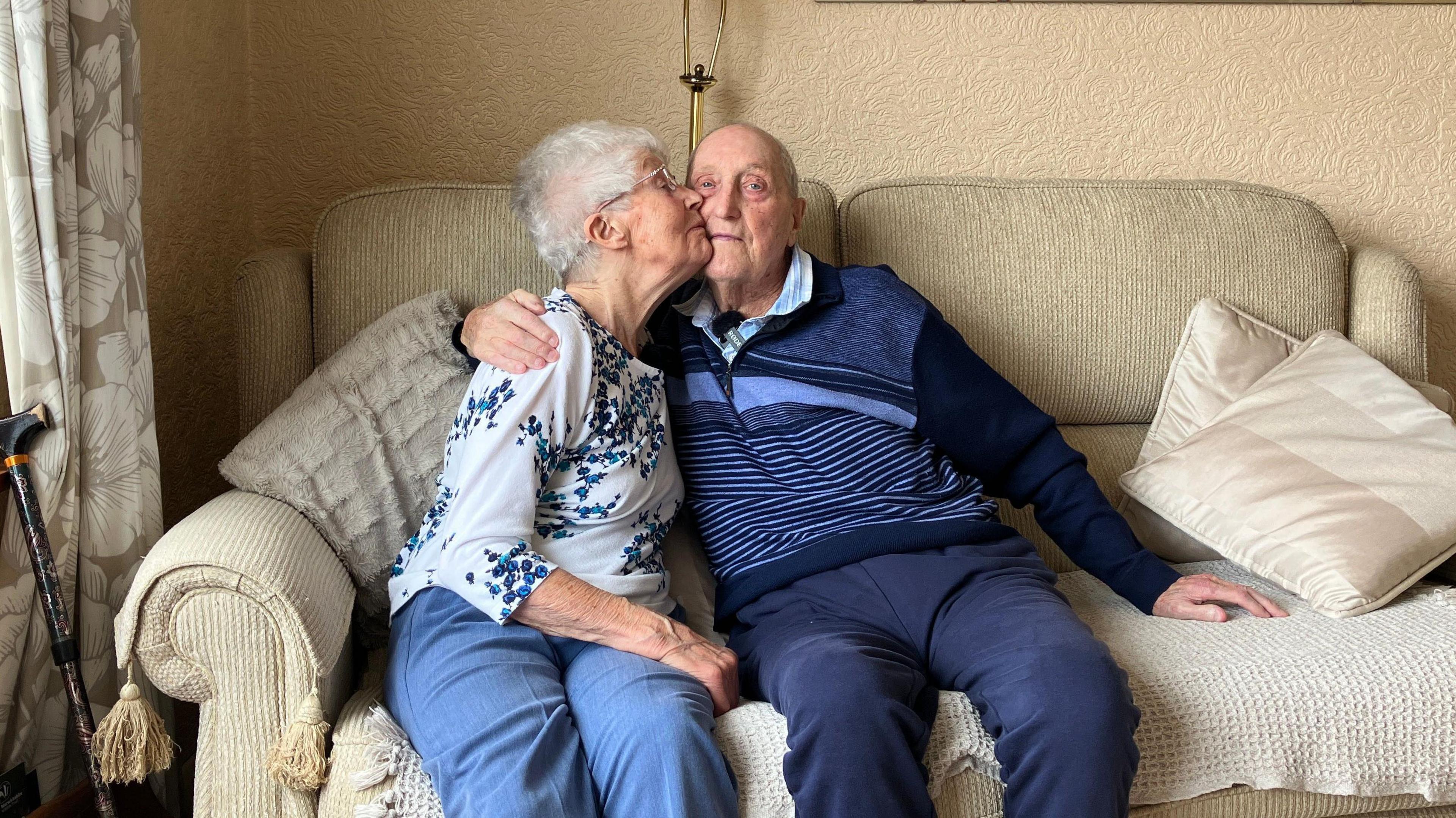 Bob and Pat Page sat on a sofa in a living room. He has his arm around her and she is placing a kiss on his cheek.