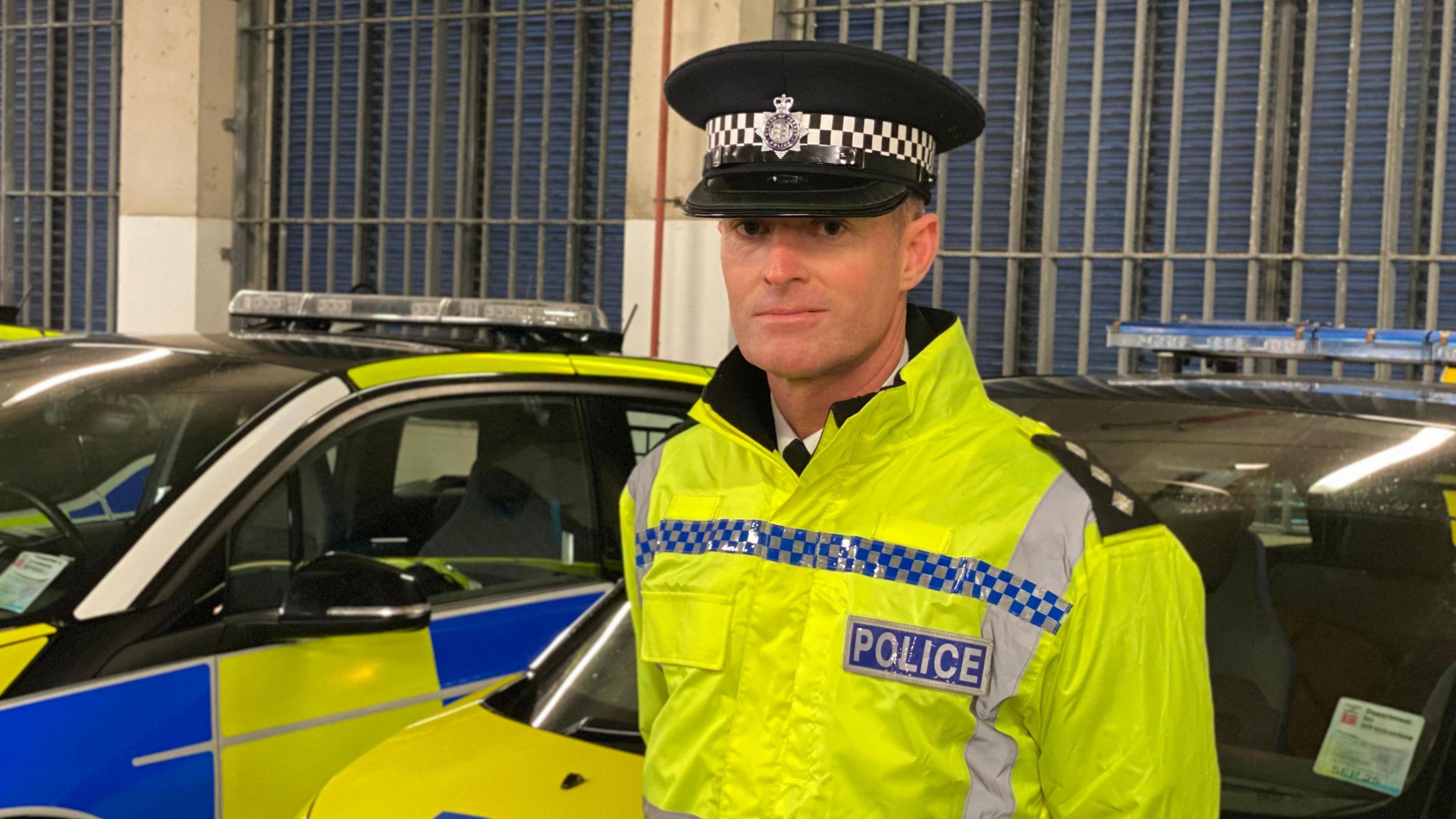 Ch Insp Mark Hafey stands in front of two police cars in a car park while wearing a yellow high-vis jacket with Police written on it. He is also wearing a flat-topped black and white police hat.