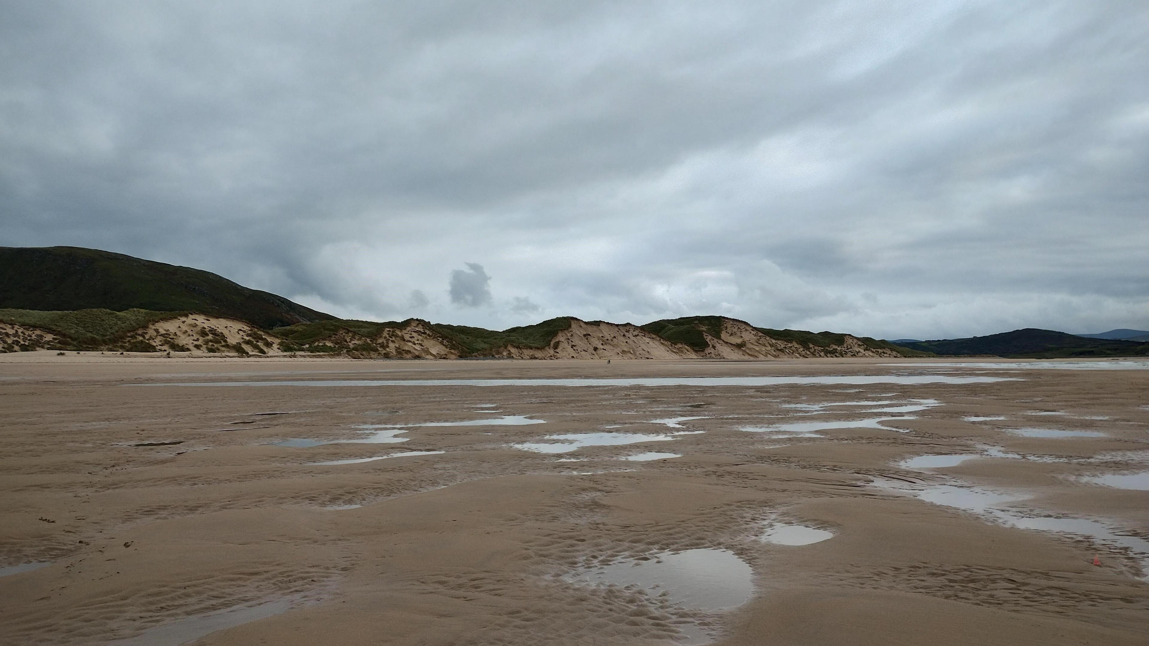 A beach on the north coast of Northern Ireland on a gloomy, cloudy day. The sky is grey/blue and there is puddles of water lying on the sand. The green sand dunes hug the skyline. 