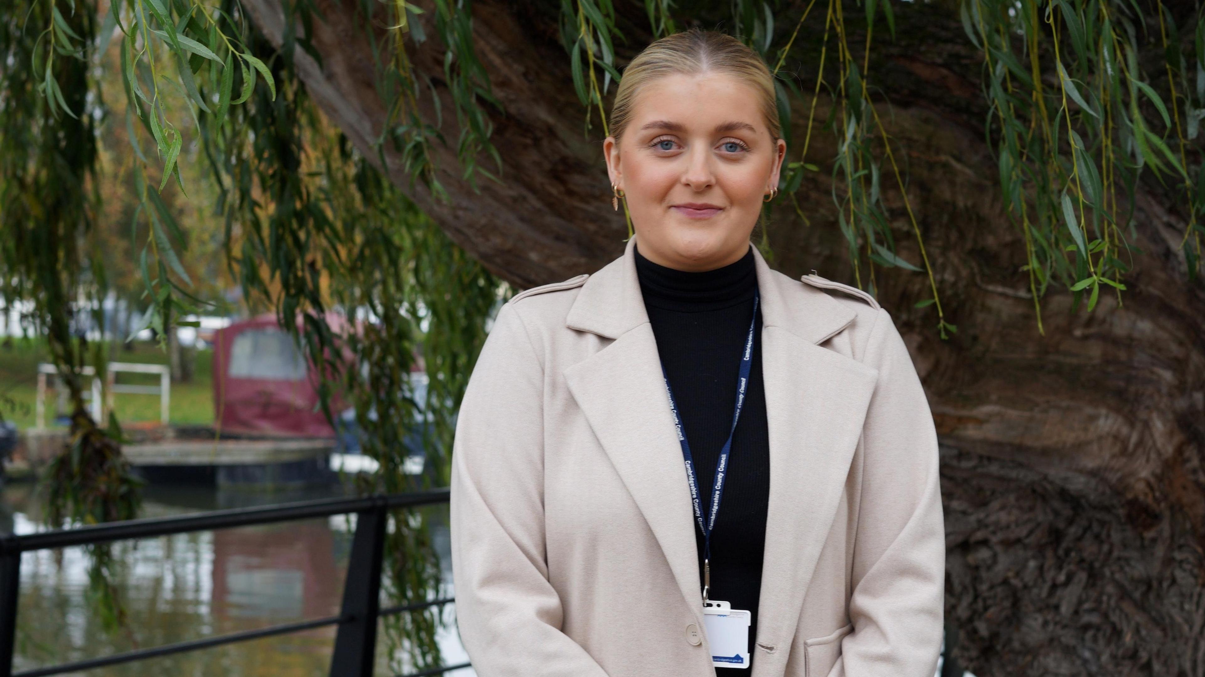 Elle Johnson is looking at the camera with a faint smile, she has a pale jacket on and black turtleneck jumper, and is wearing a lanyard. She is standing in front of a weeping willow beside the canal in Ely.