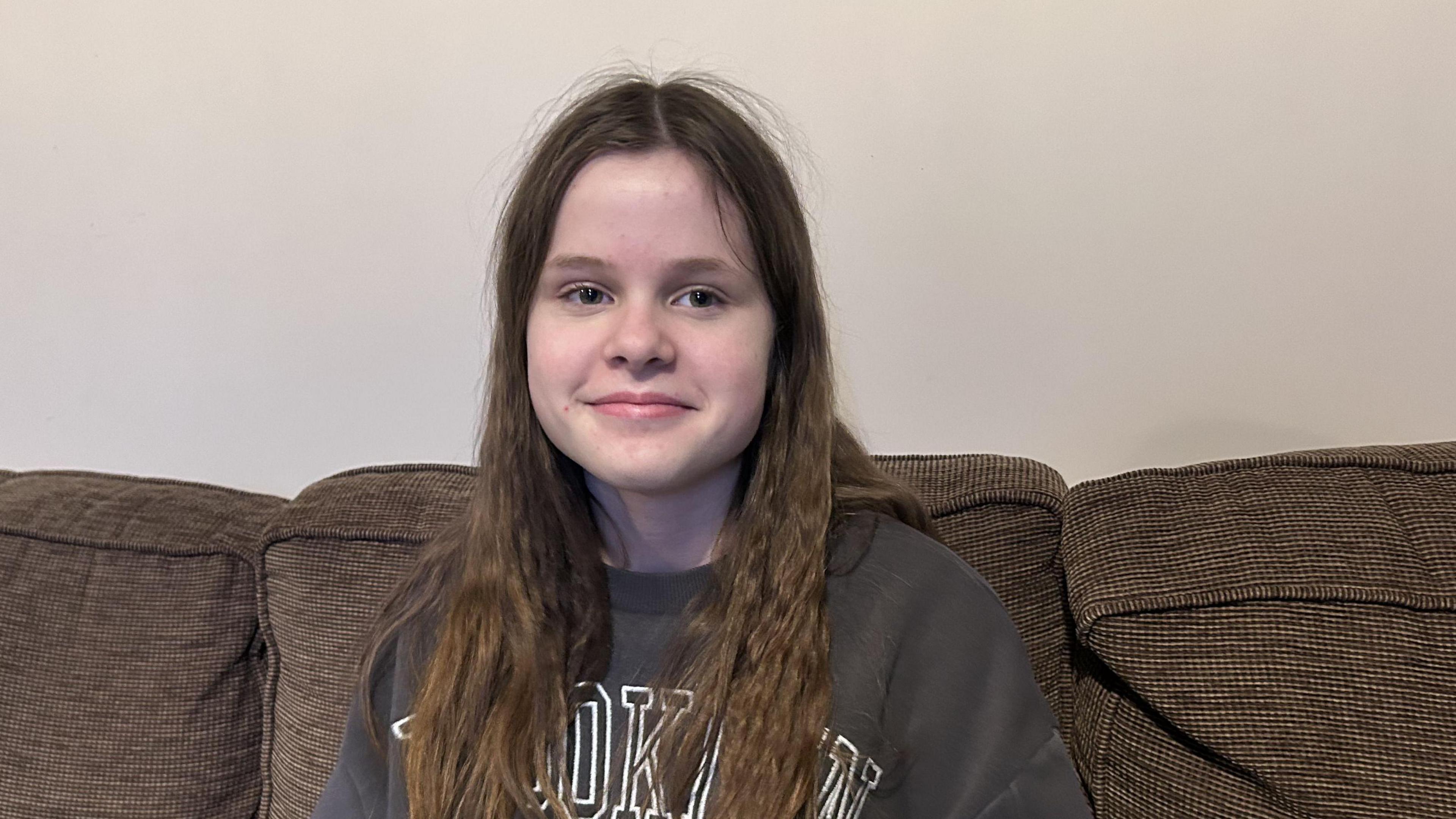 Lucy  with long wavy dark hair sits smiling on a brown sofa. She is wearing a black sweatshirt.