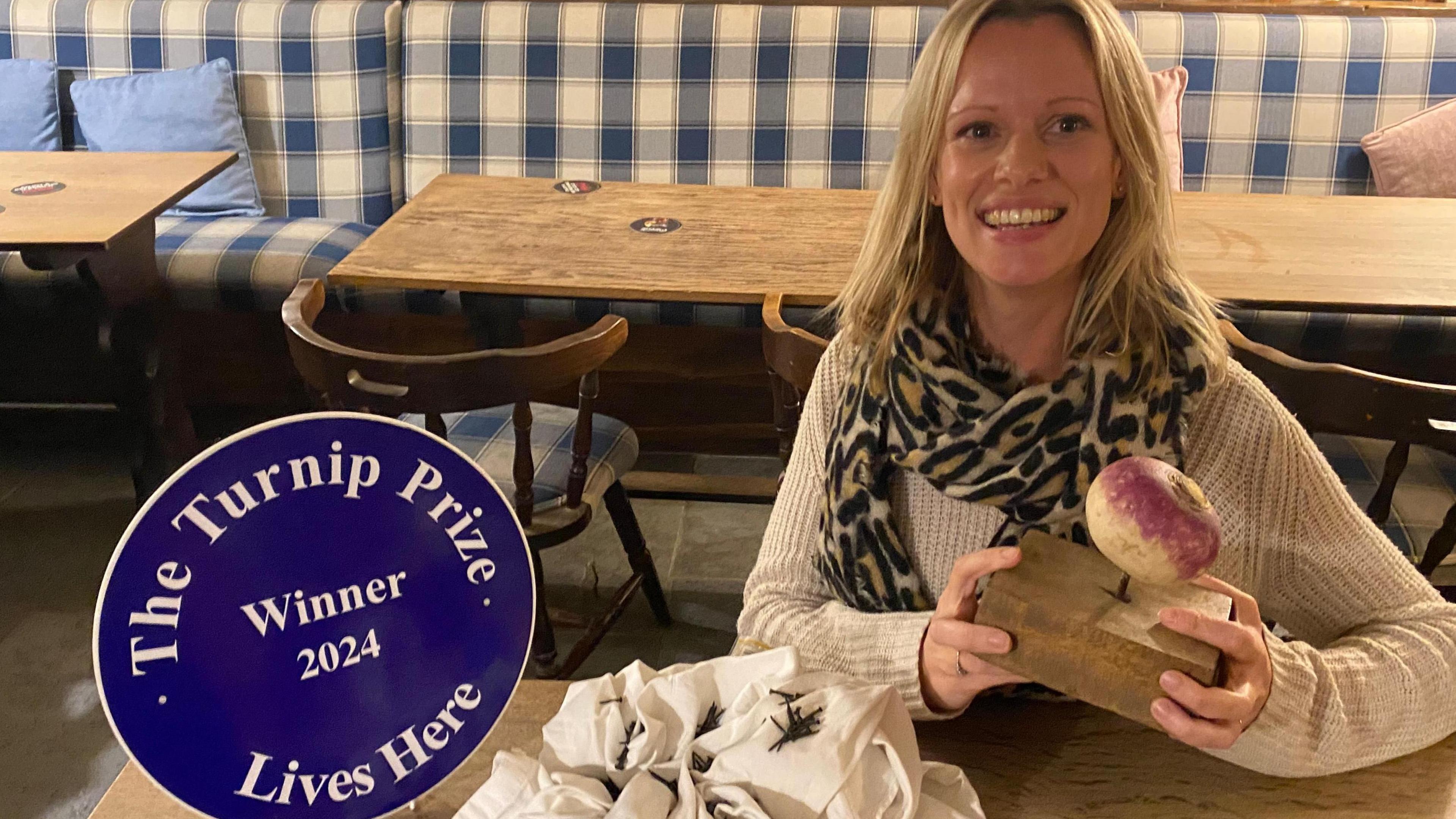 A woman smiles at the camera holding a turnip with a nail through it. She is sitting next to a sign saying 'Turnip Prize winner' and a white crumpled up shirt with tacks in it.