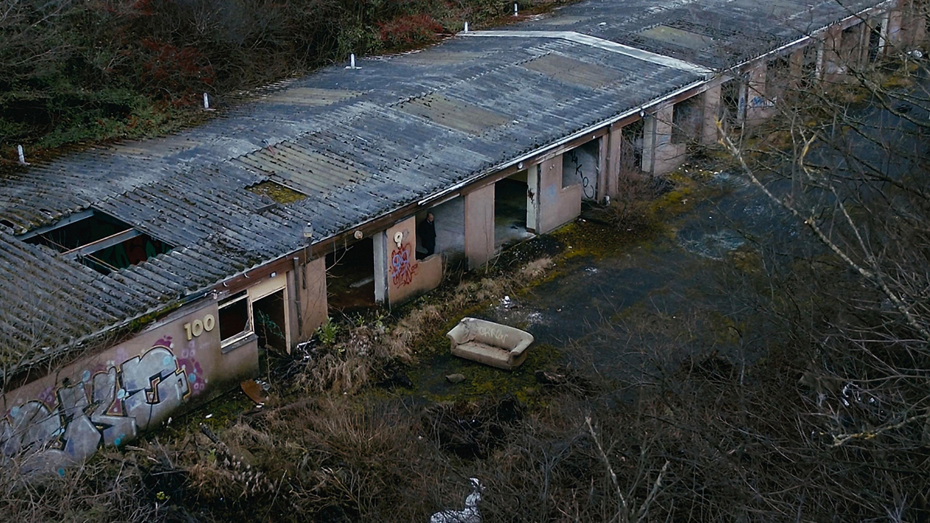 An abandoned warehouse with lots of individual units. There are holes in the roof and the building has no windows or doors. It has been spray painted with graffiti and there is an old sofa outside 