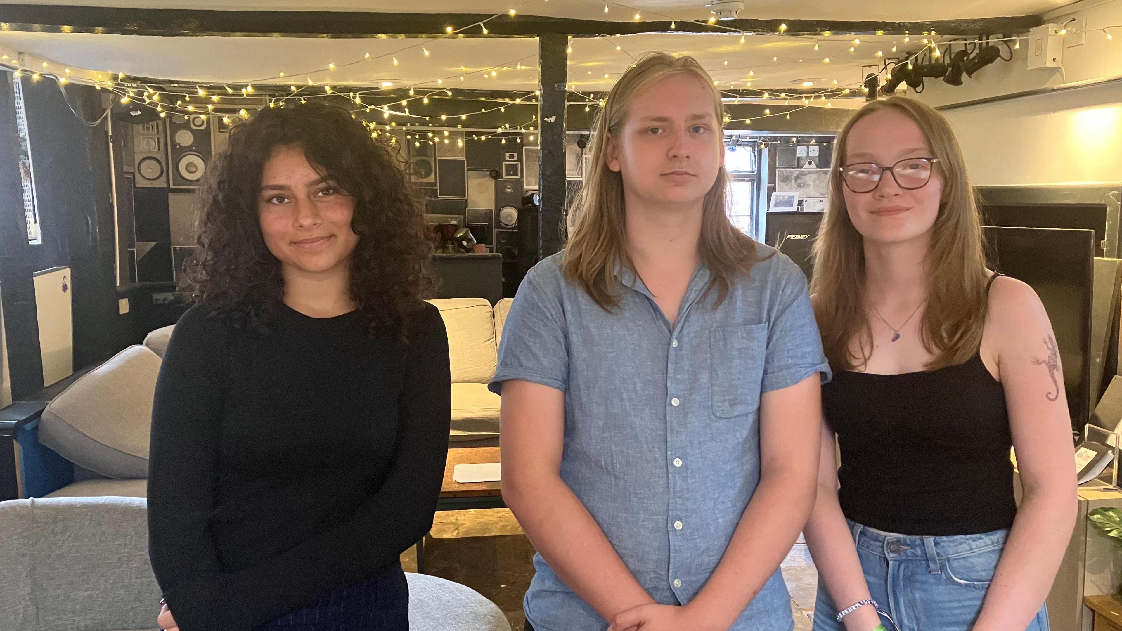 A picture of three teenagers, aged 16 and 17, stood in a room. There are fairy lights on the ceiling behind them. On the left is a young woman, with dark shoulder length curly hair who has a black jumper on. In the middle a young man, with long blonde hair who has a blue short sleeved shirt on. On the right is a young woman with dark blonde hair, glasses, and a black top on, and you can see she has blue jeans on.