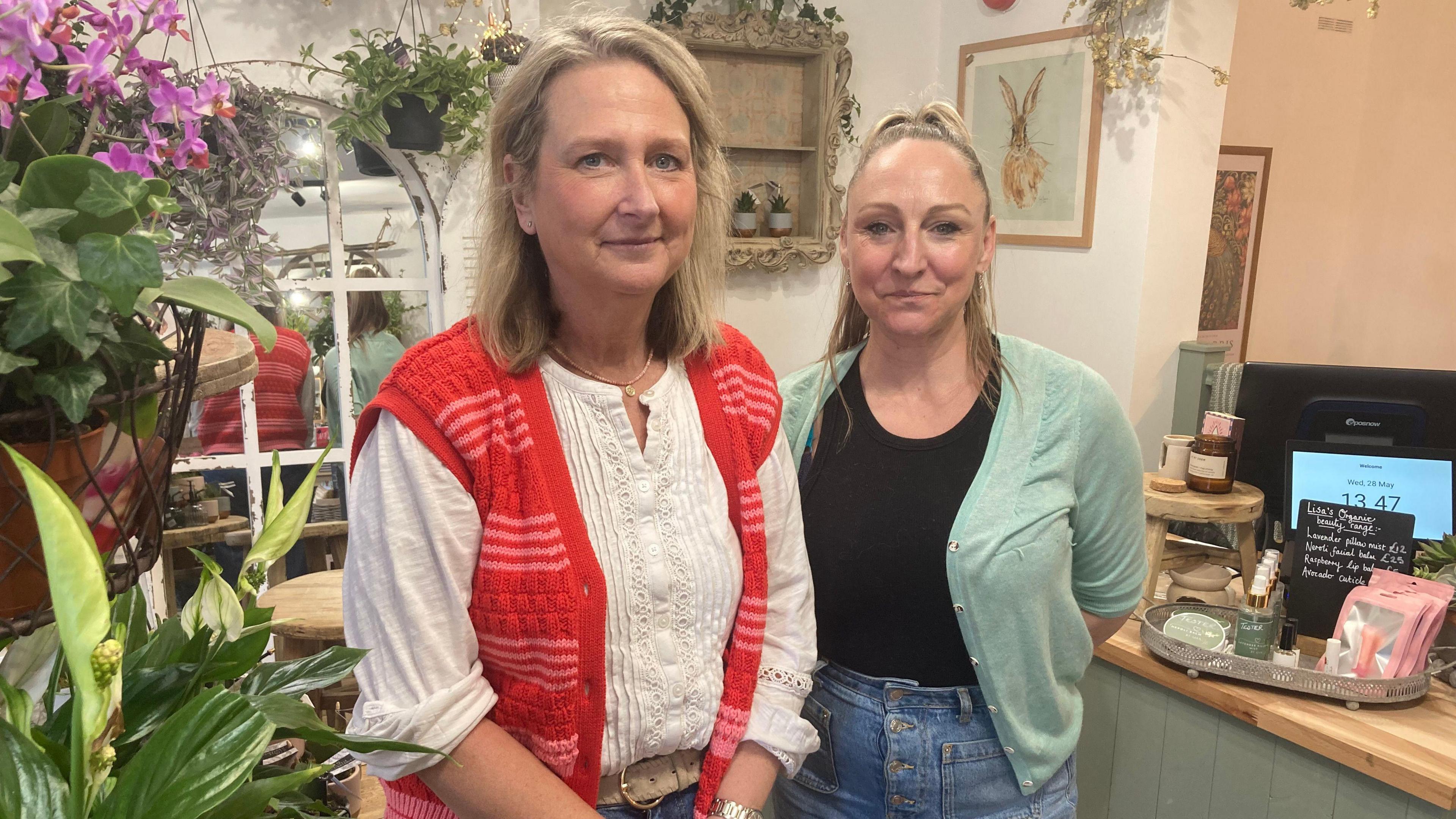 Carly Johnson and Lisa Foster. They are standing in their shop. Both of them are blonde. Carly is wearing a white blouse and a red and pink gilet. Lisa is wearing a black t-shirt and a mint green cardigan. Both of them are smiling.
