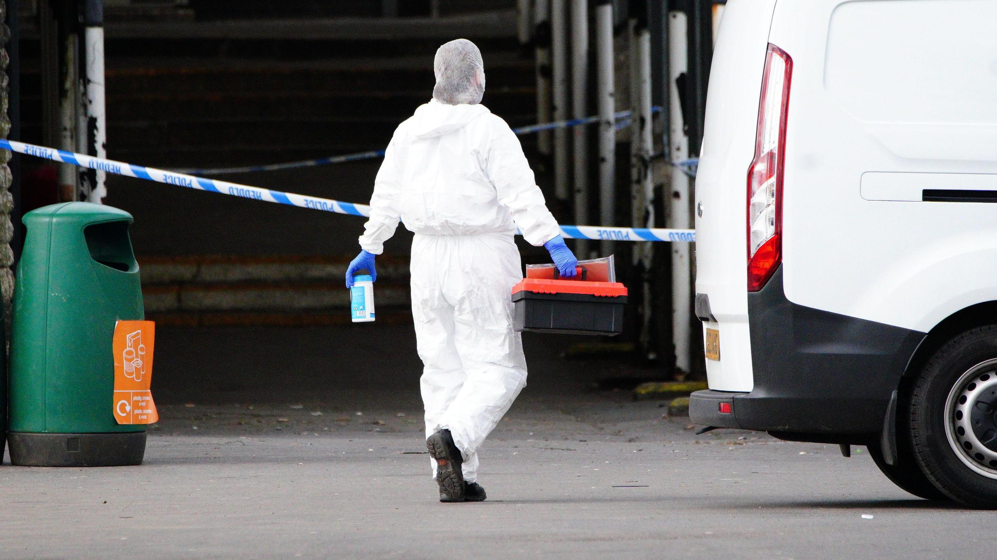 A forensic investigator at Amman Valley school, in Ammanford, Carmarthenshire, on Wednesday, April 24, 2024. To their right is a white van and to their left a green bin. 
The person is walking toward a line of police tape