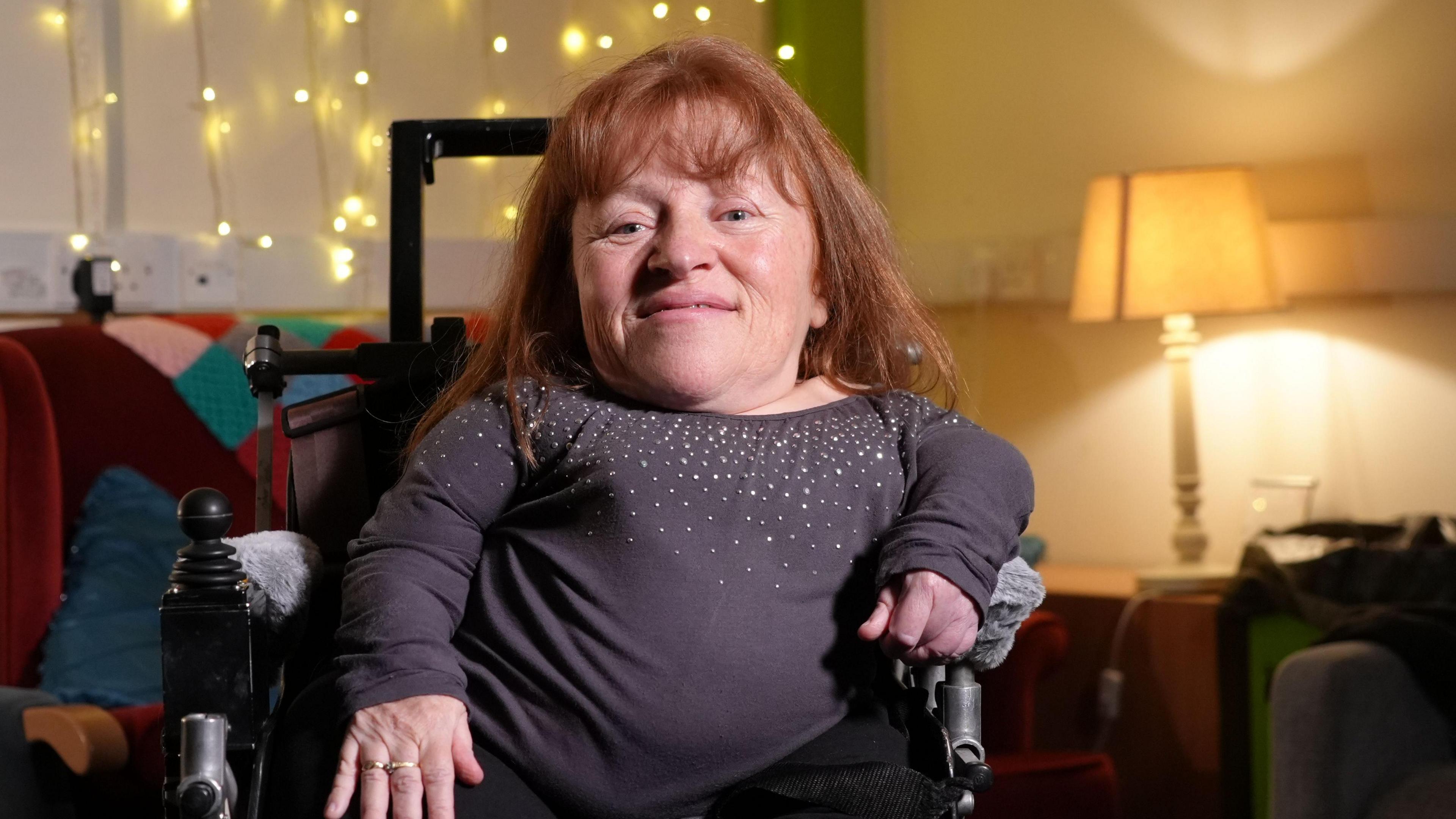 Angie Stewart is in her wheelchair and looking at the camera. She has long ginger hair and is wearing a purple top with sparkles on it.