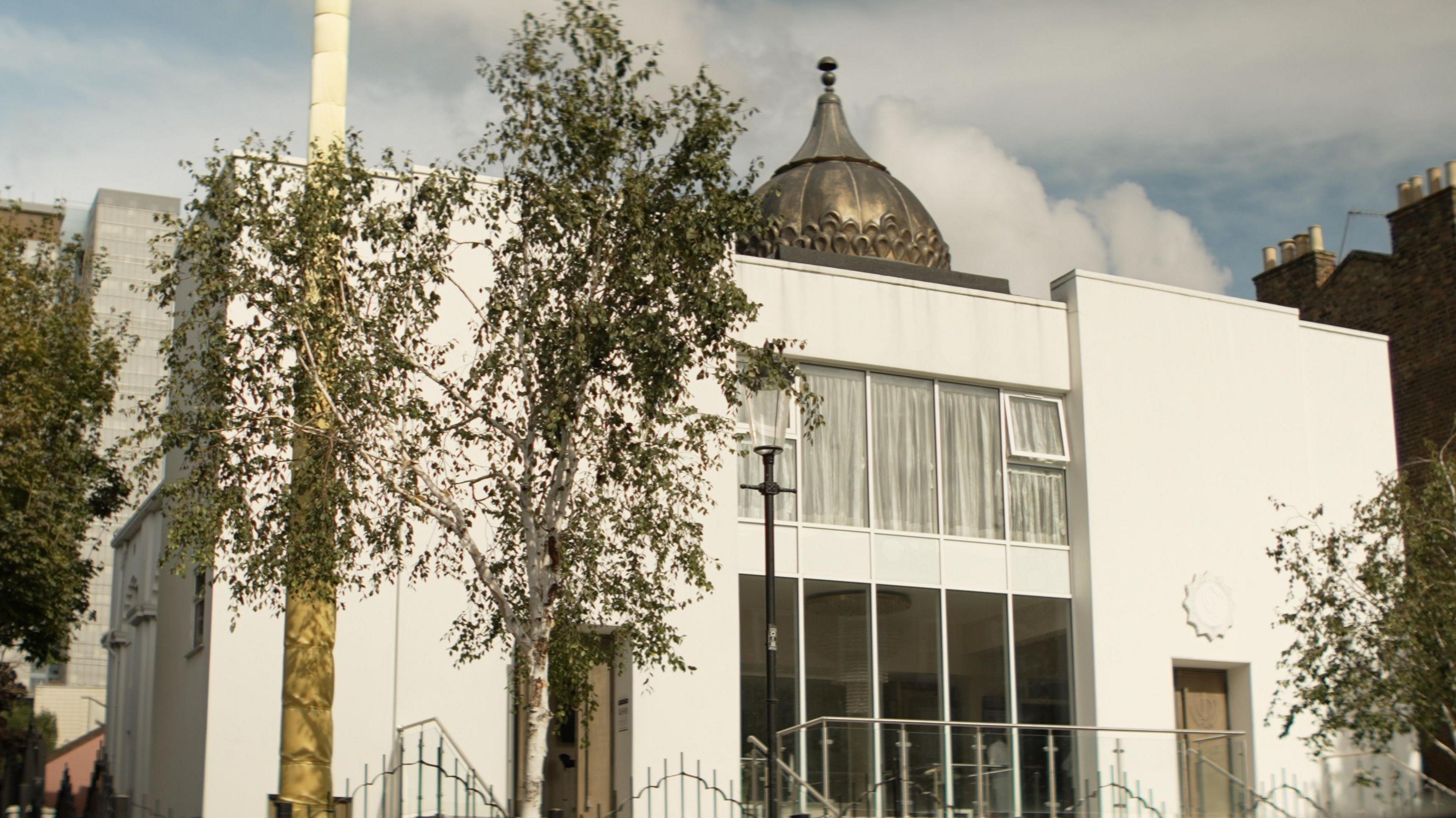 A large white building with silver metal railings at the entrance and small silver birch saplings framing the entrance. On the roof there is a copper colour decorative minaret.
