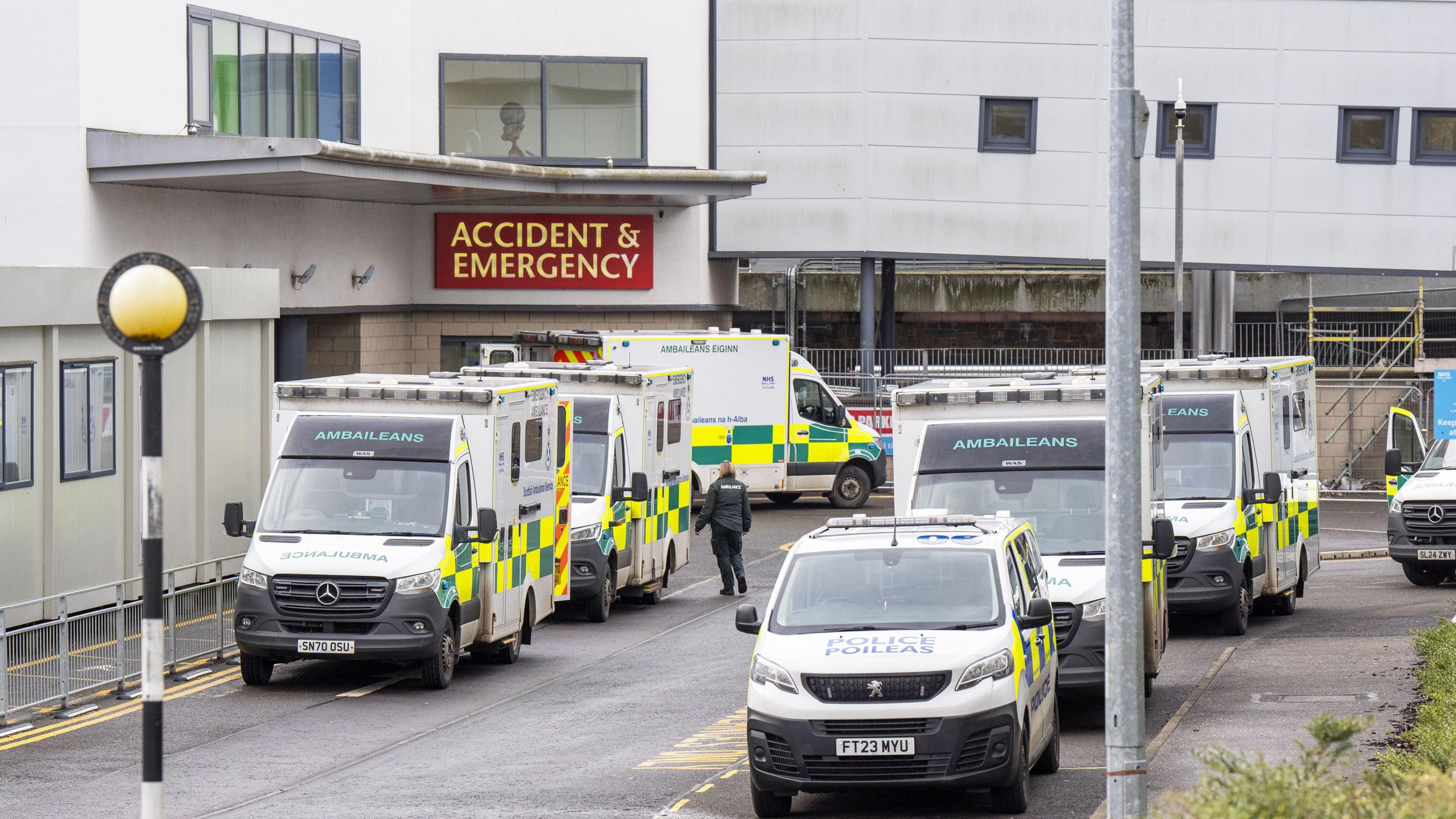 A hospital building, with an ACCIDENT & EMERGENCY sign above the doors. There are several ambulances, a police van, and paramedics outside the doors.