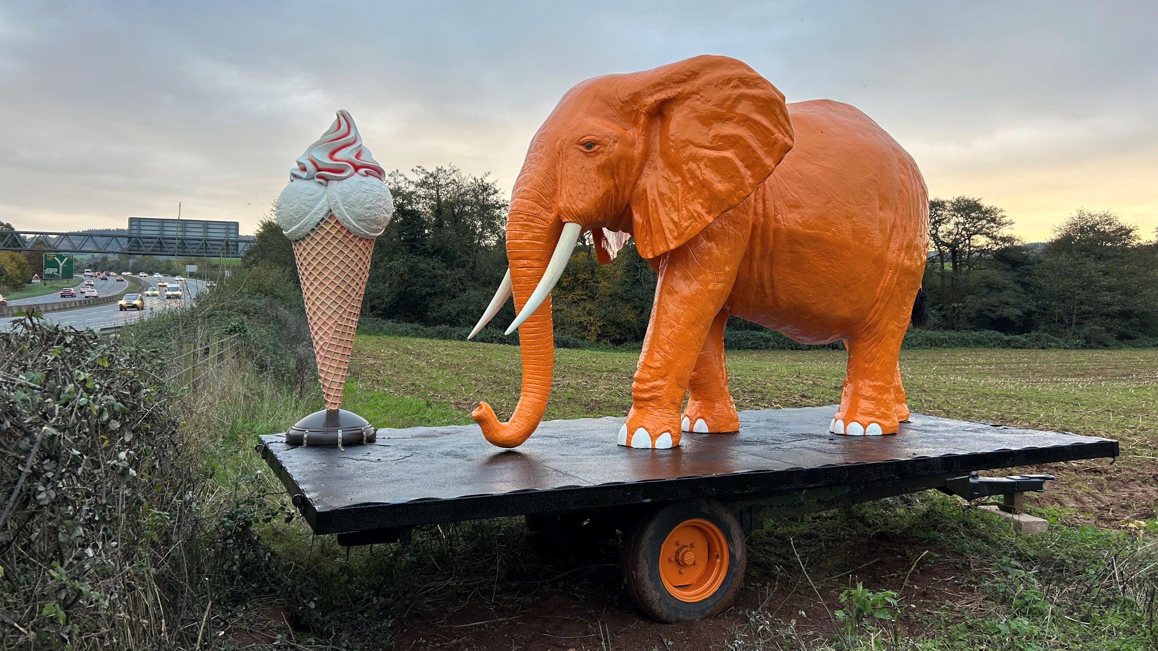 A giant ice cream cone statue next to a large orange elephant statue stood in a field on a Devon farm - on a black wooden base which is on wheels. Cars travelling on the A38 dual carriageway are visible on the left of the field the statue is in.