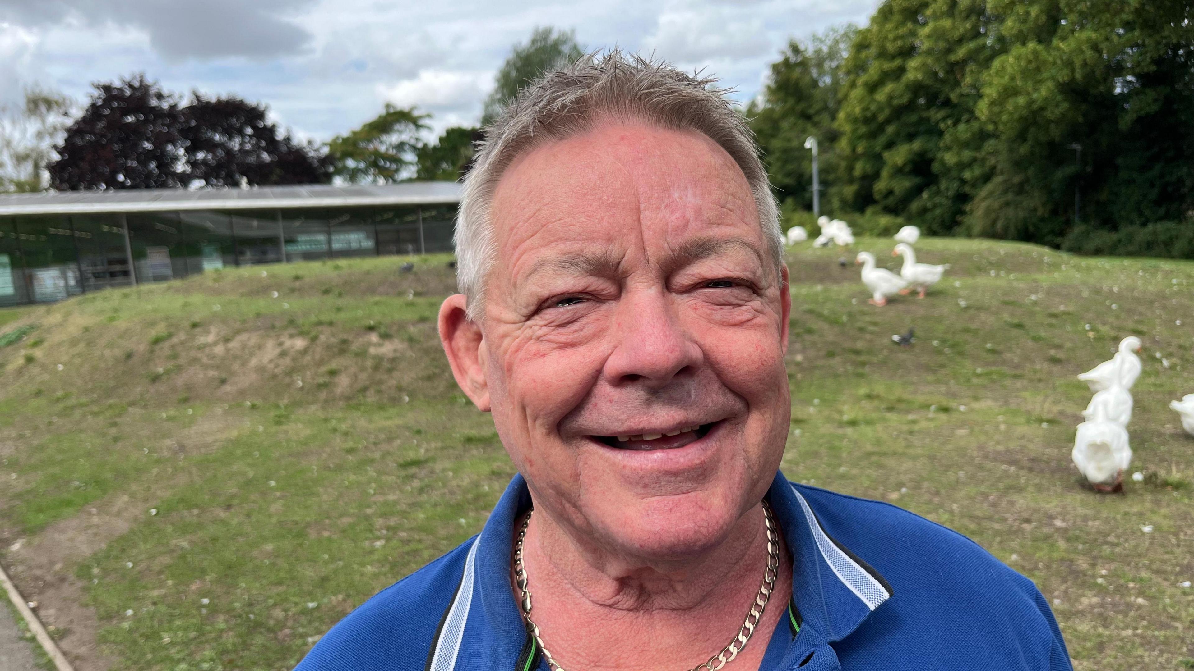 Kevin McCourt, 72, has short greyish hair, he is wearing a blue open necked T-shirt with a thick gold chain underneath. Behind him are some white geese on a small hillock.