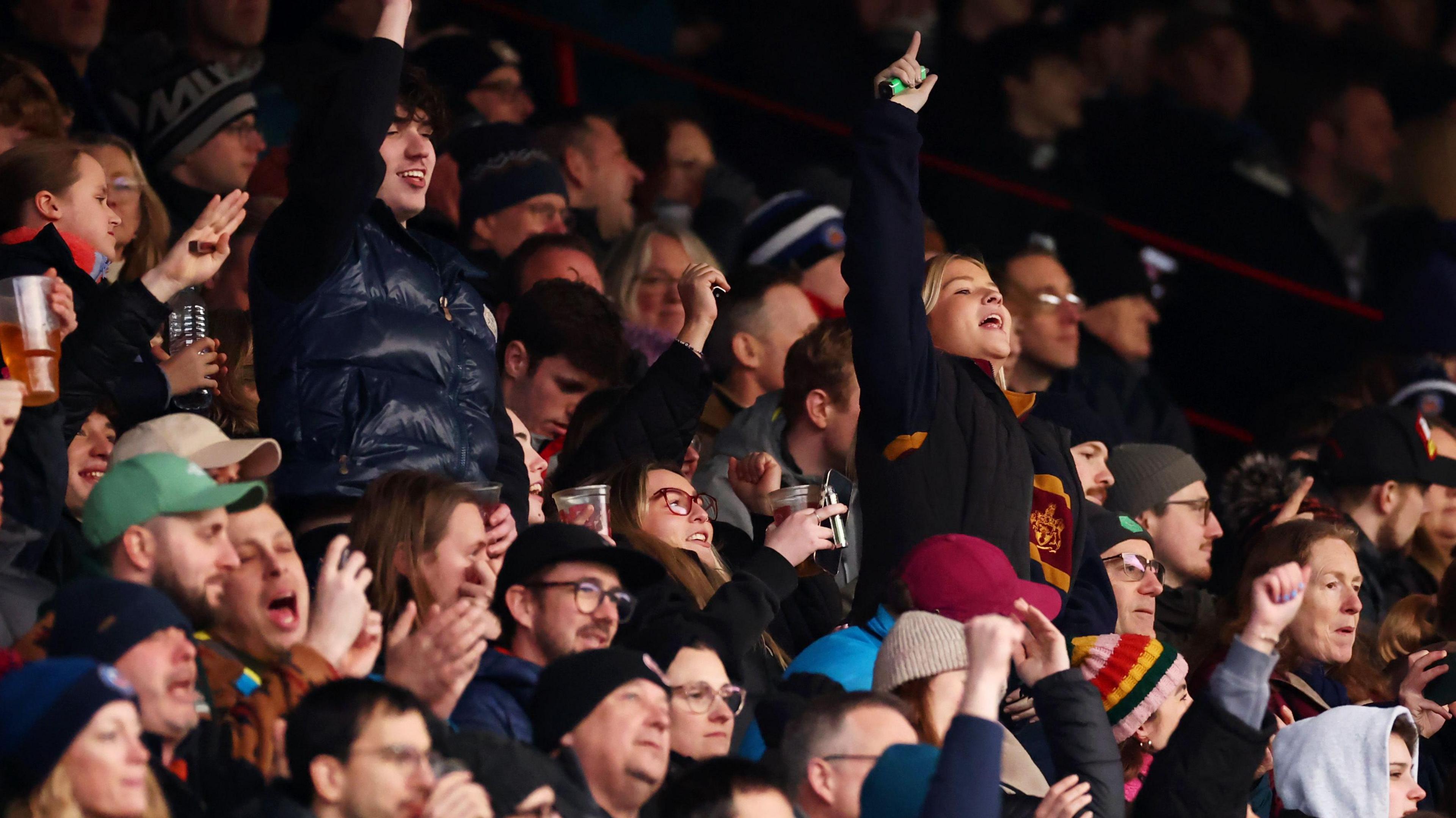 A stand full of rugby fans, some of them standing and holding their arms aloft, cheer on the teams as Bristol play Bath at Ashton Gate