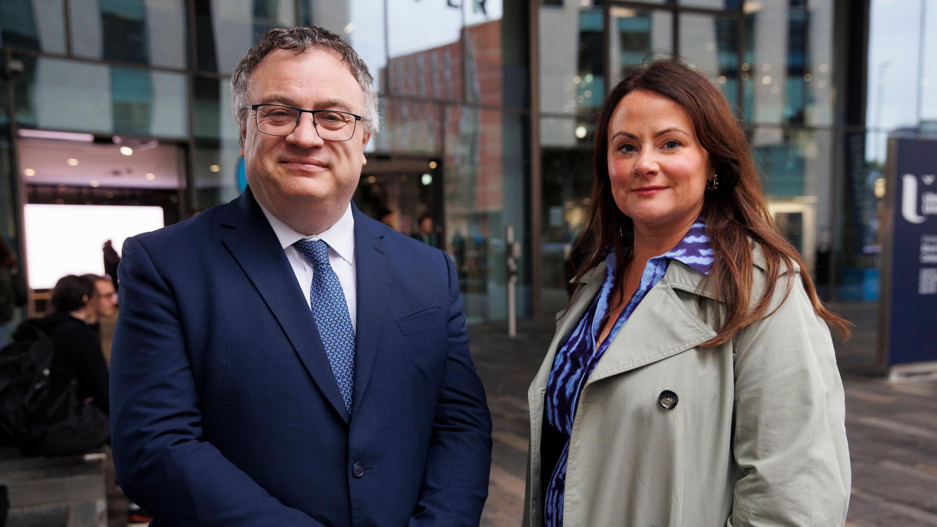 Former Alliance deputy leader Stephen Farry and former Stormont special adviser Jodie Carson. Farry on the left is smiling and wearing a blue suit, white shirt and blue tie. He has short grey-black hair and black-rimmed glasses. Carson is wearing a blue dress and grey mac. She has shoulder-length brown hair.