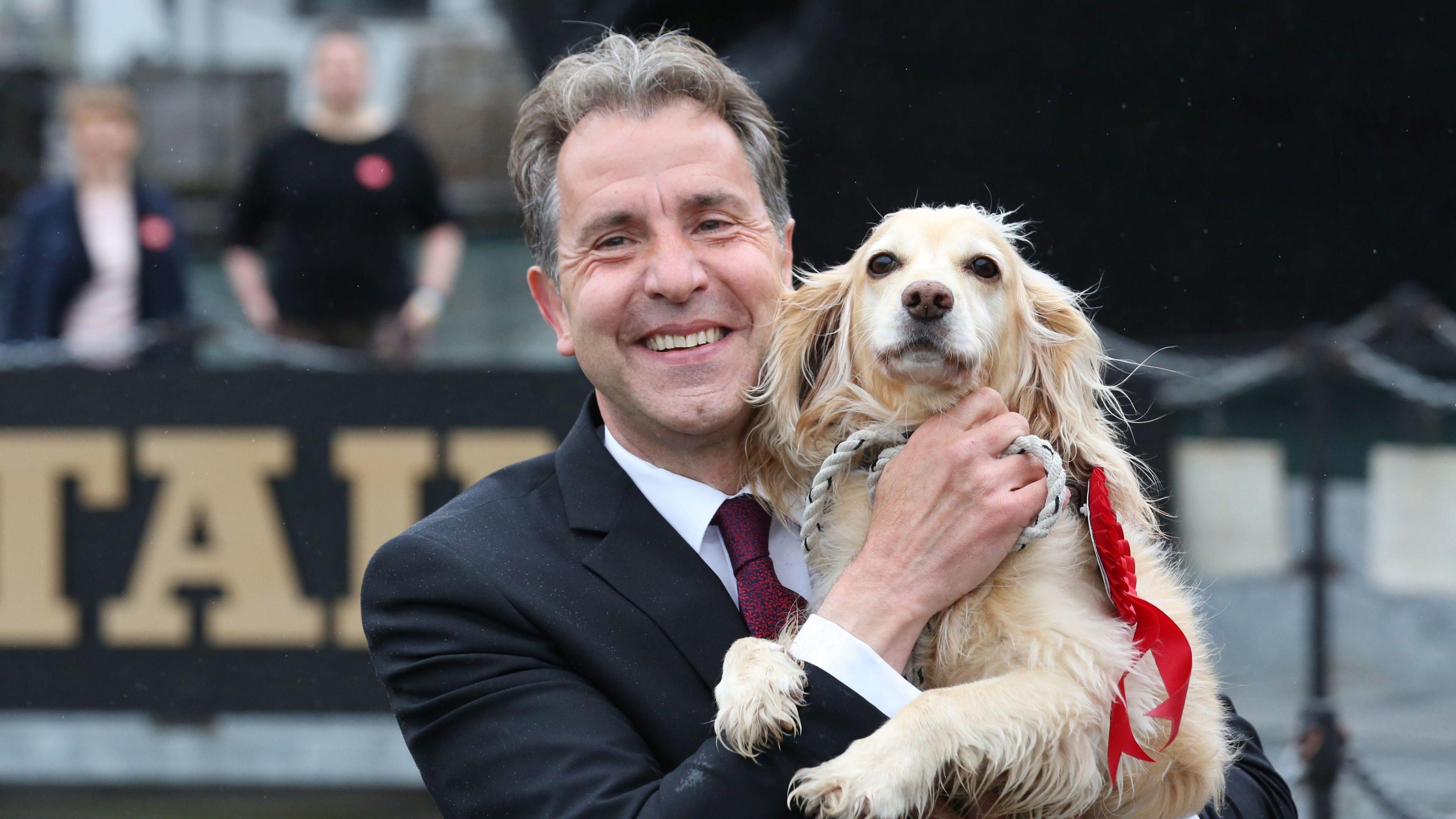 Metro Mayor and MP Dan Norris smiles at the camera while holding up his golden spaniel dog who wears a red rosette 