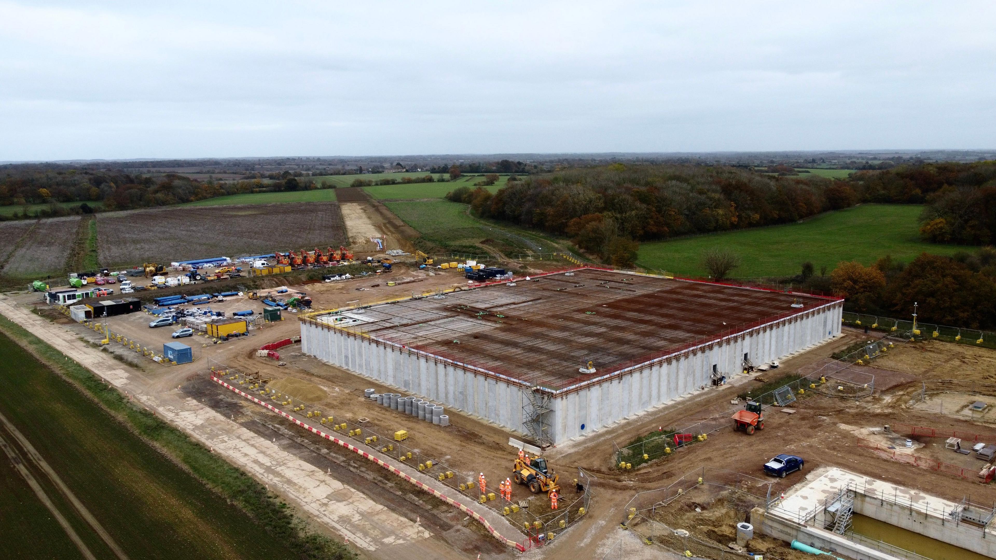 A drone image of a development site. A large square building is in the process of being built. Workers in orange hi-vis suits can be seen working around the site. Other building infrastructure can seen around the square building.