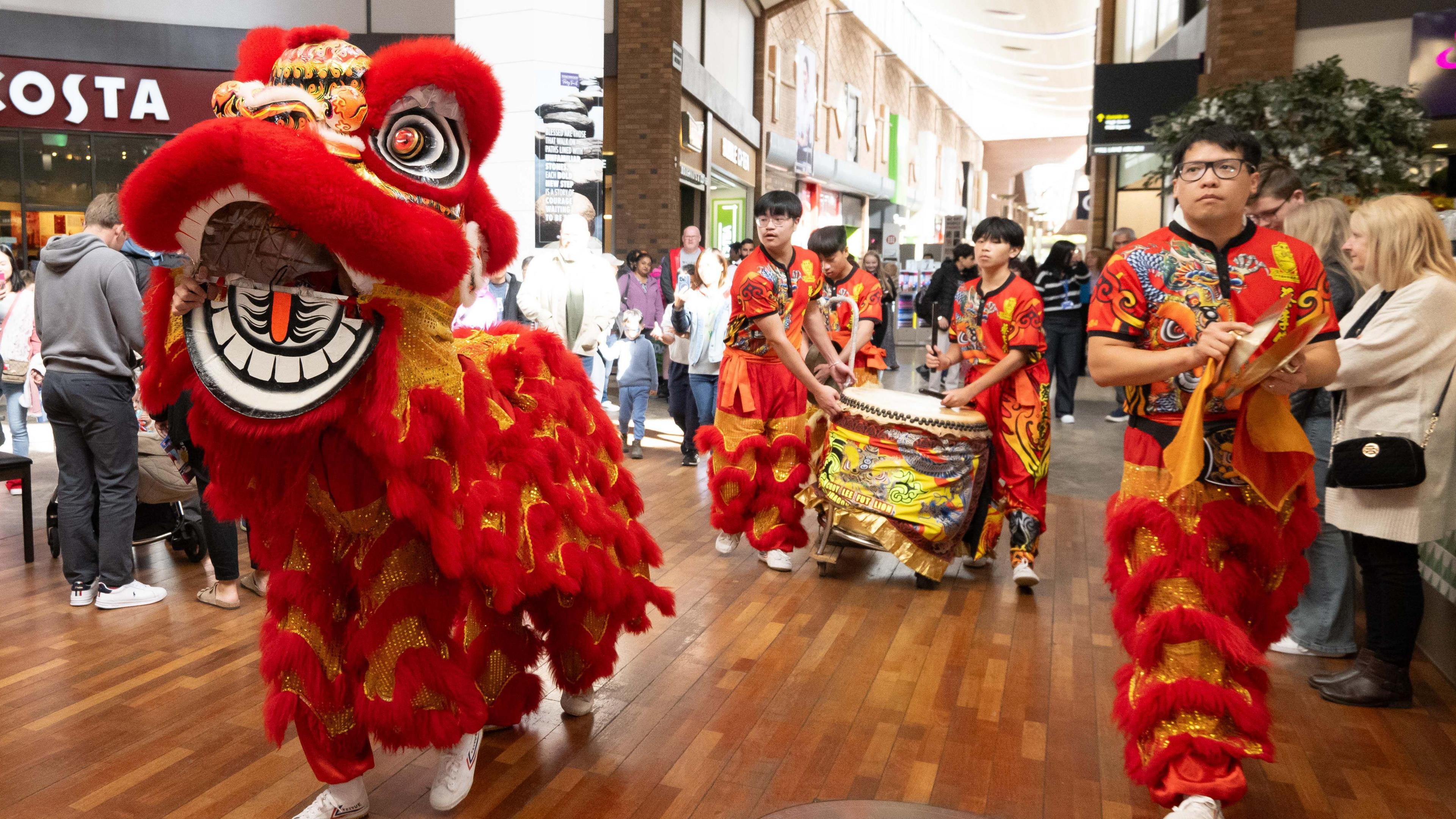 Two performers in a Chinese dragon costume and four other men in bright red and orange and orange decorated festival costumes walk through the centre of a shopping centre, watched by crowds. Three of the men are carrying a large drum, wrapped in a yellow-patterned cloth.