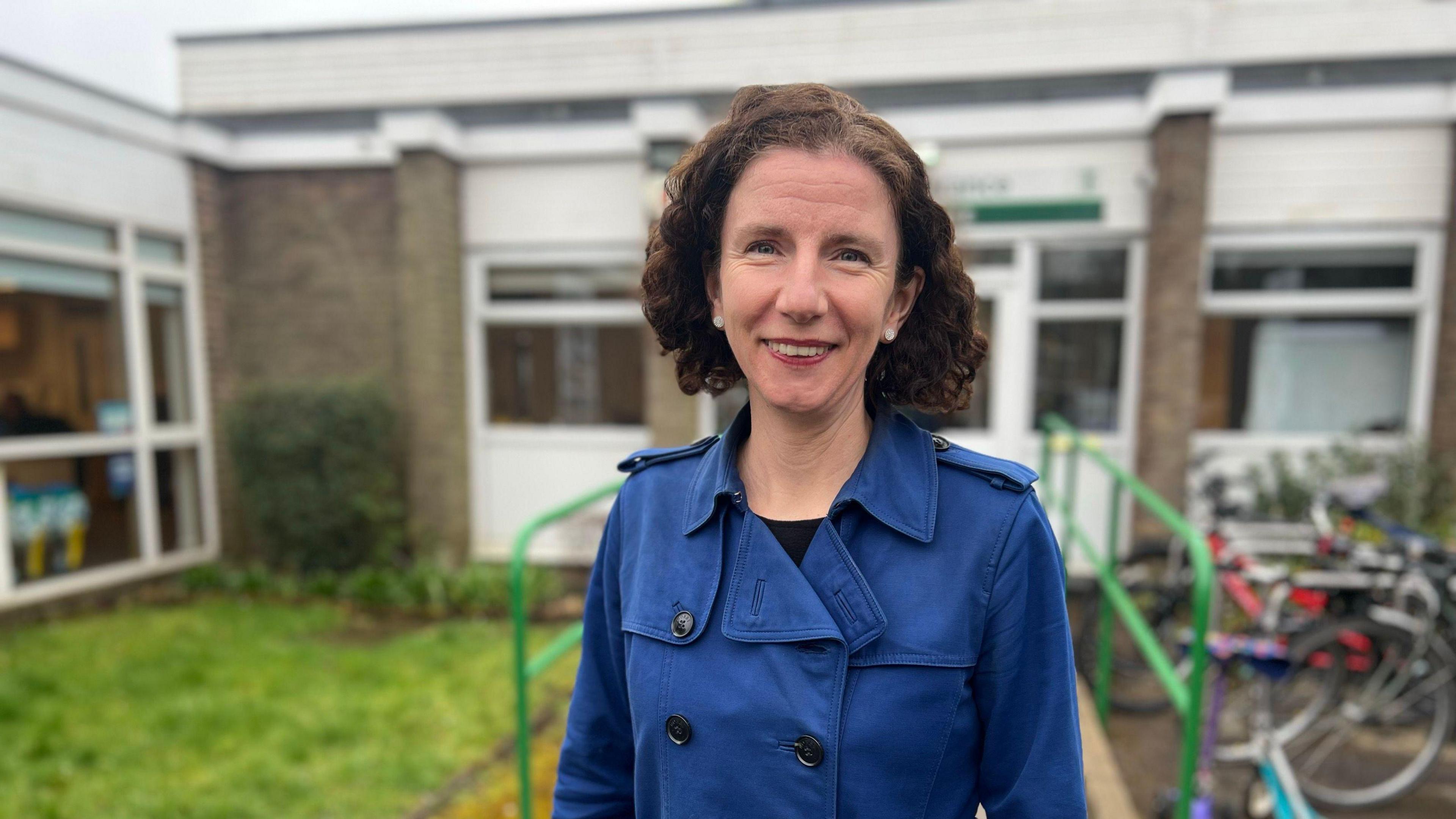 Anneliese Dodds stands in front of a school in a blue coat. She has dark hair, almost at shoulder length.