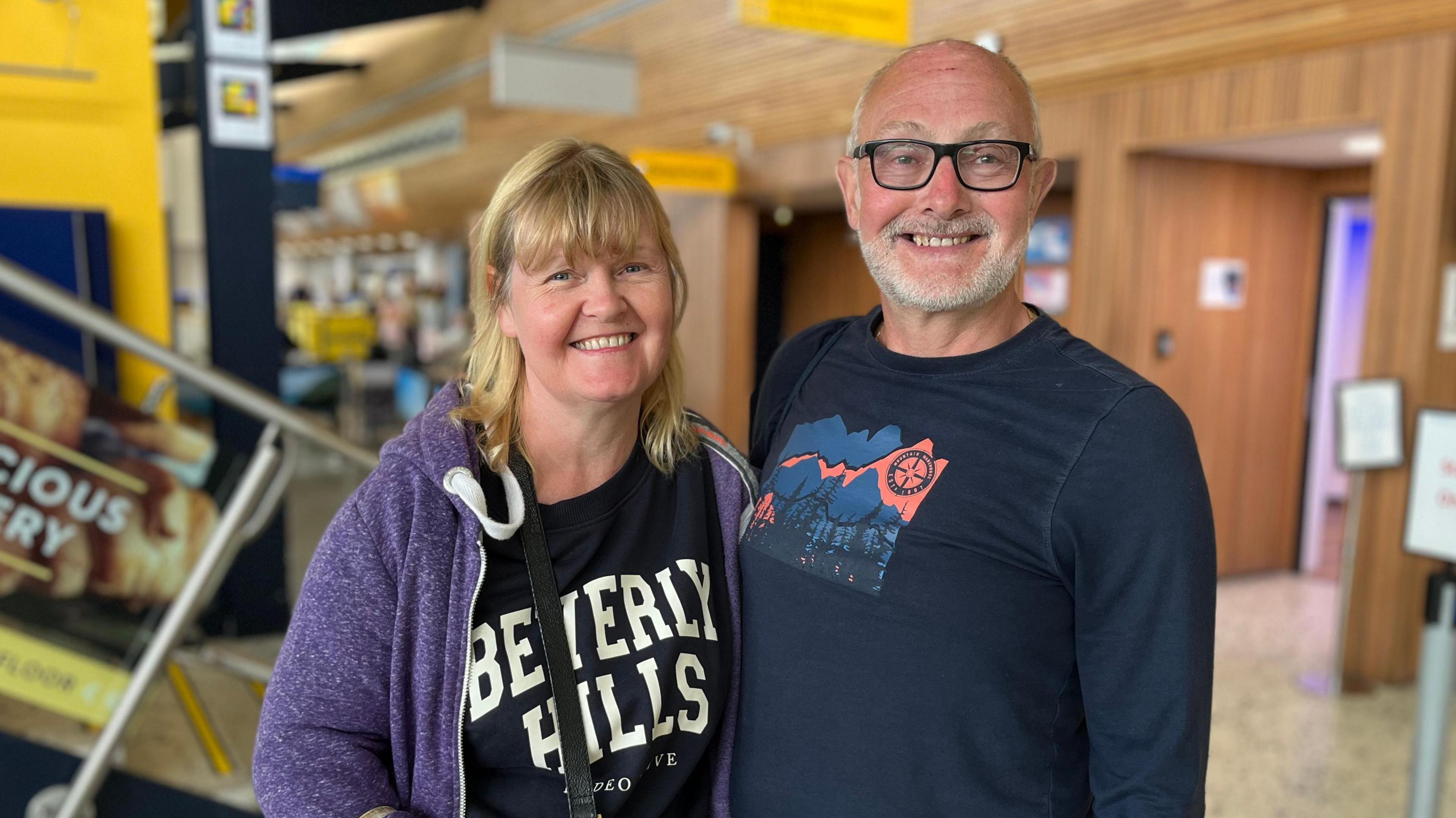 Dominique has shoulder length blonde hair and is wearing a black t-shirt with a purple zipped jumper over the top. John is weraing black framed glasses and a black long sleeve t-shirt. They are both looking at the camera and smiling.