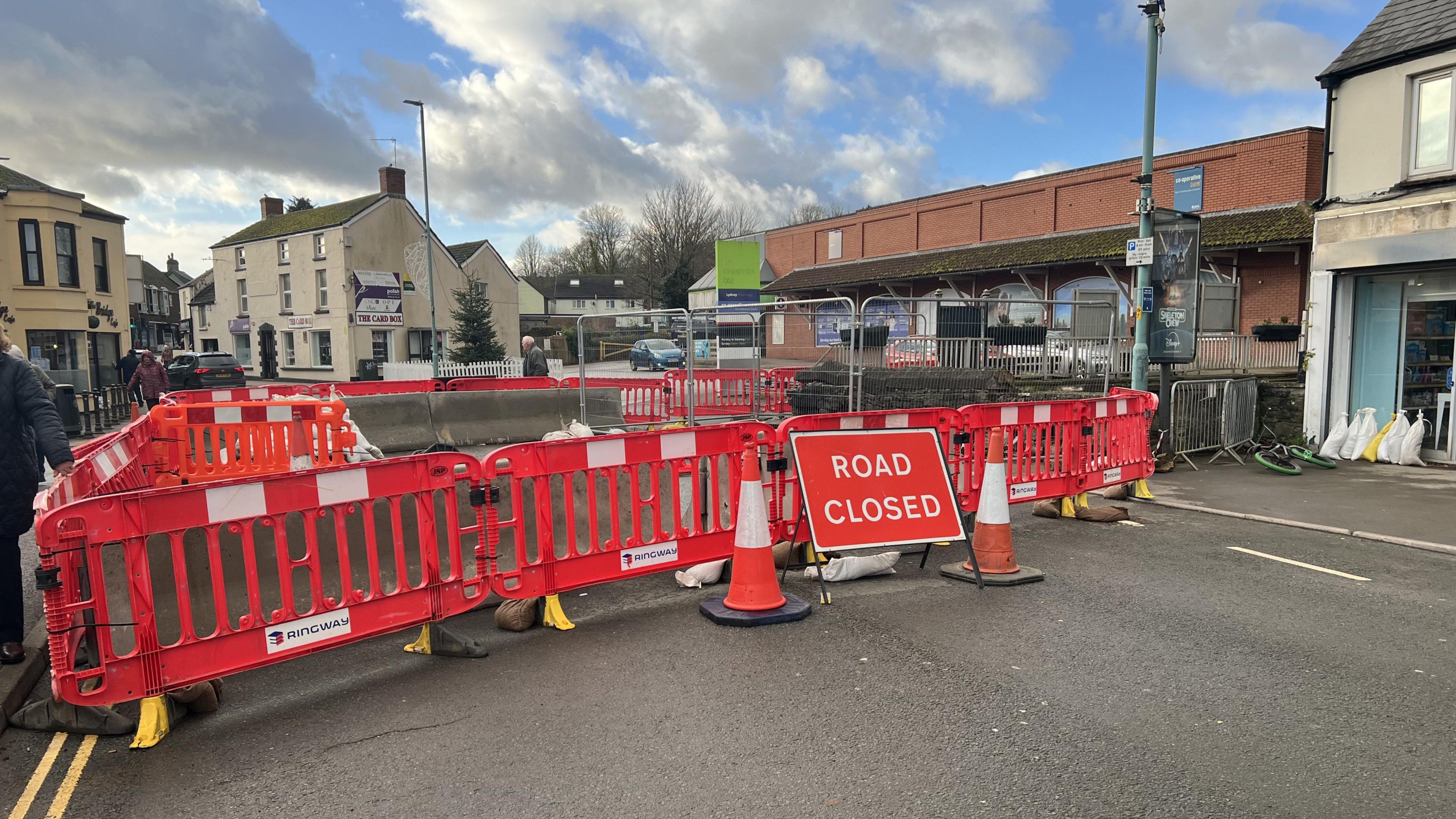 A large perimeter of red barriers blocking the road from any vehicles. There are also several concrete barriers as reinforcement. There are orange cones and a road closed sign. The whole street is blocked off and only pedestrians can walk along the pavement on the left. The damaged bridge is on the right behind metal fencing.