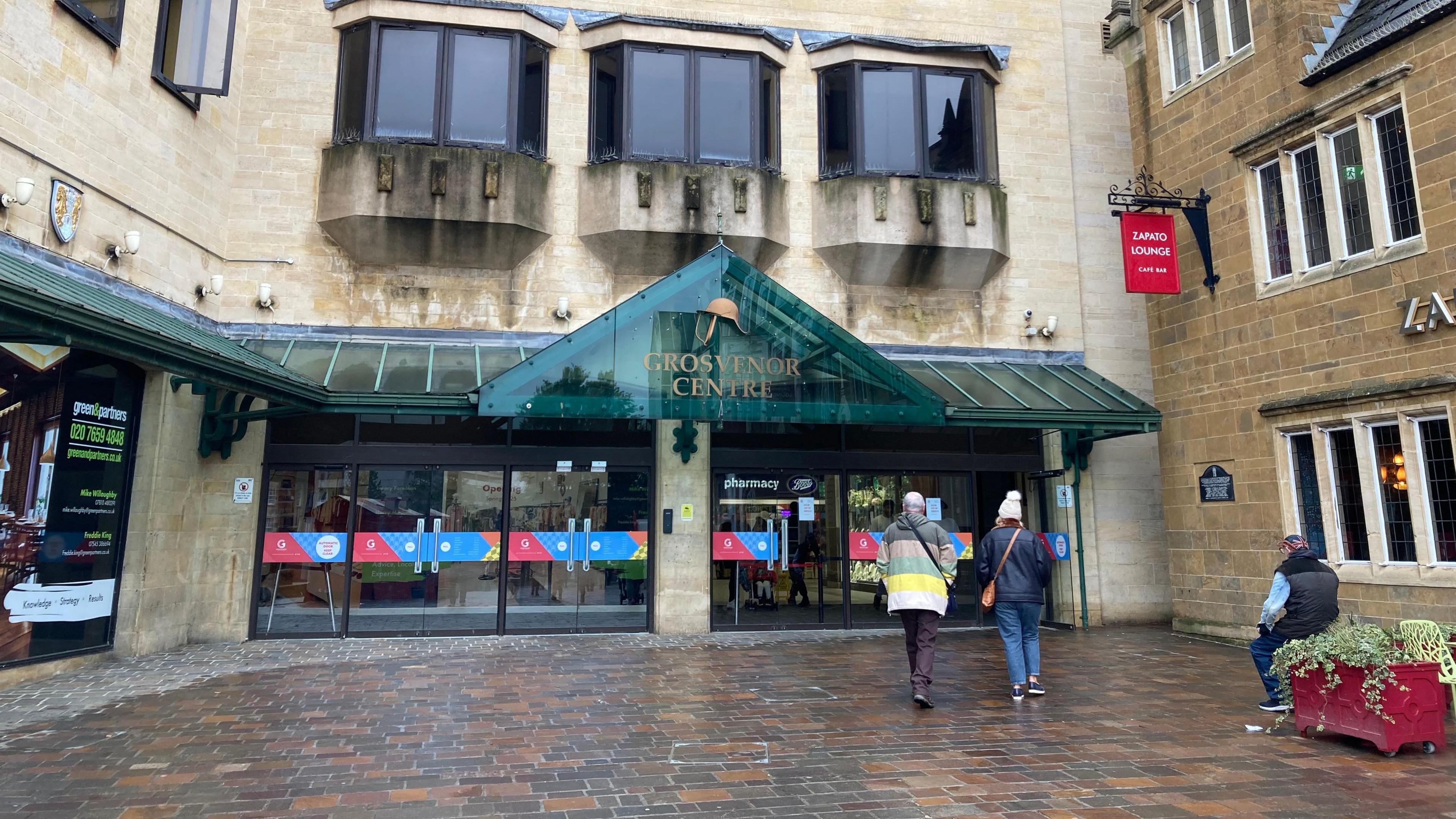 A glass frontage with eight double glass doors and a green glass rain cover displaying the sign for the Grosvenor Centre.