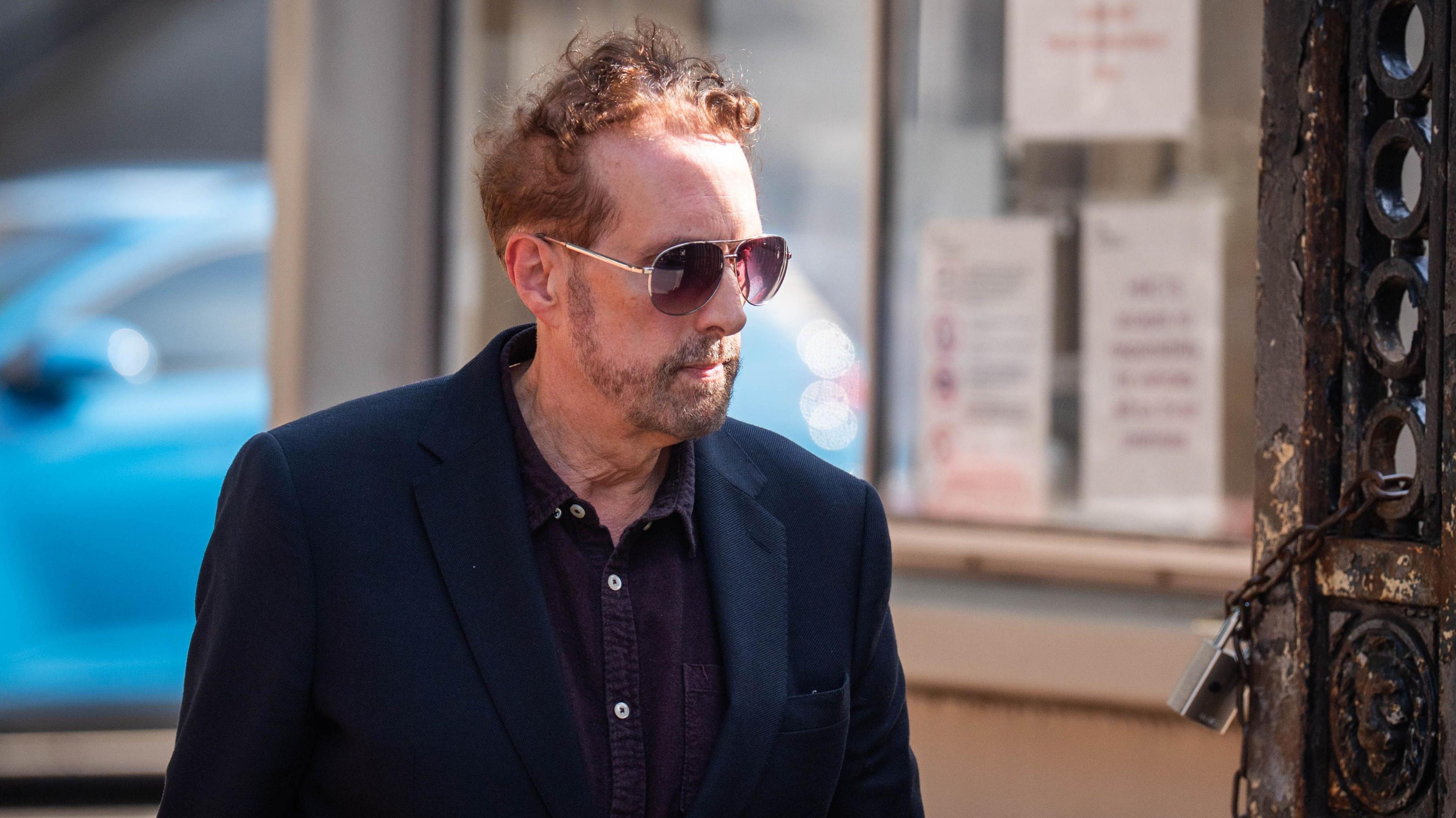The image depicts a Chris Brain walking outdoors during the day. He is wearing a dark blazer and sunglasses, and has short hair along with facial hair. In the background, there is a blue car parked near a building that has several notices or signs posted on its window.