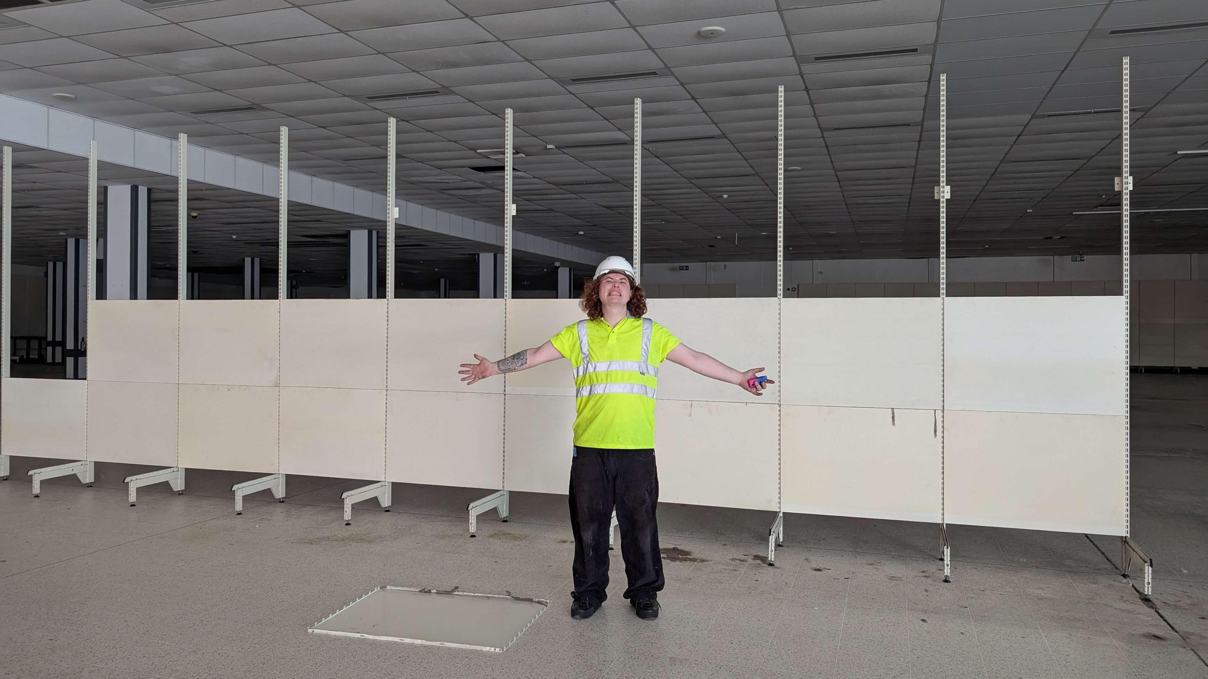 Theodore Godfrey-Cass stands inside the empty retail unit. He has shoulder-length brown hair and is wearing a hi-vis jacket and a white hard hat. Behind him are large white panels which divide the large store in two.