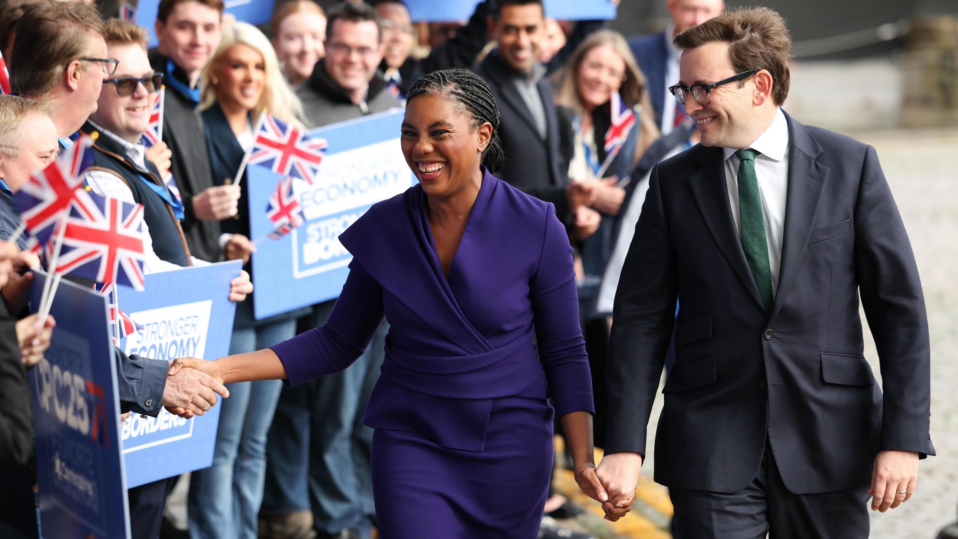 Badenoch wearing a blue top and dress and holding hands with her husband who wears a dark grey suit and green tie. They are being greeted by party supporters holding Conservative placards and waving Union jack flags.