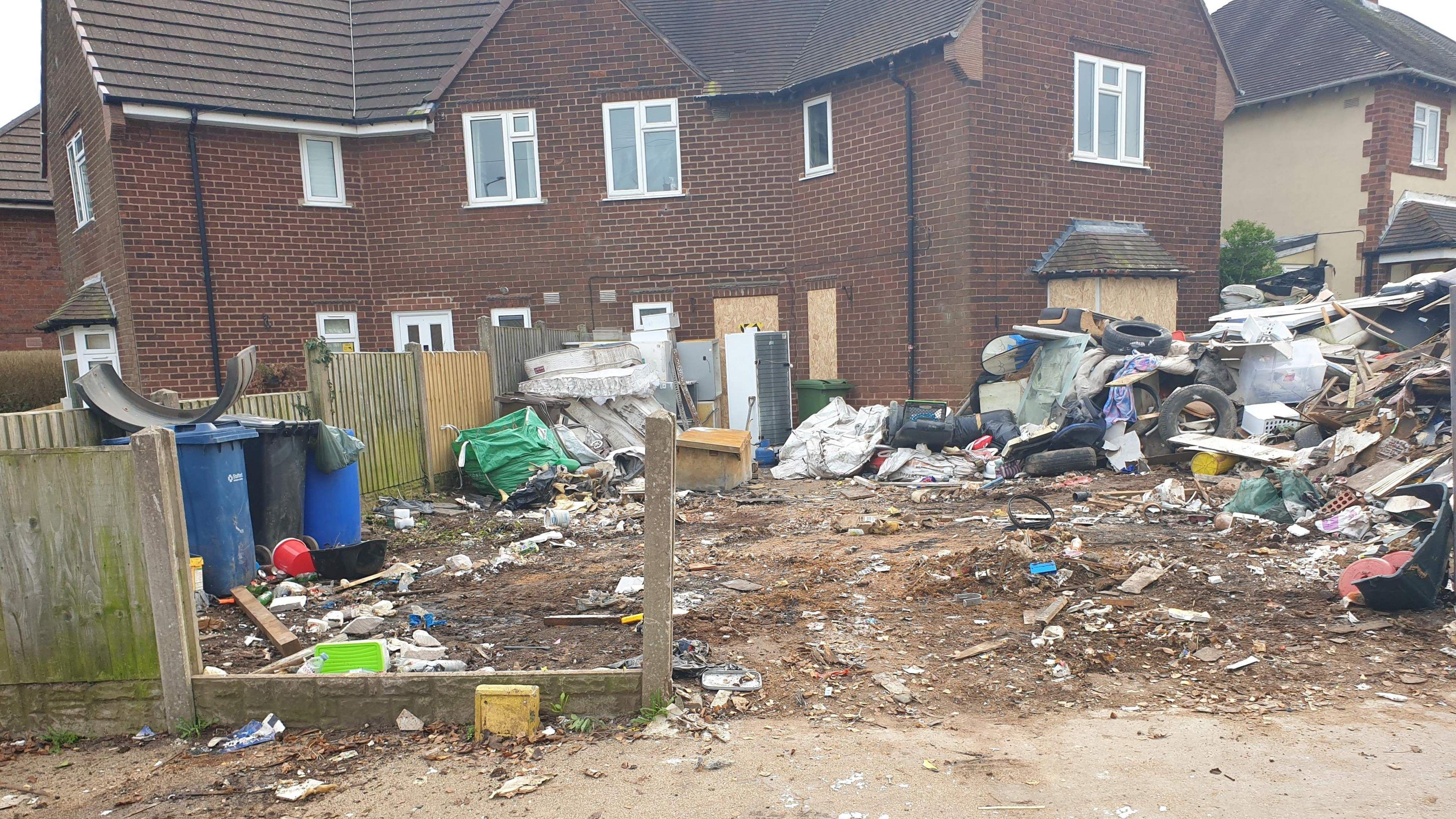 A pile of rubbish in the front garden of a house on the right with three bins on the left. Wooden panels have been placed across the windows and doors of the property.