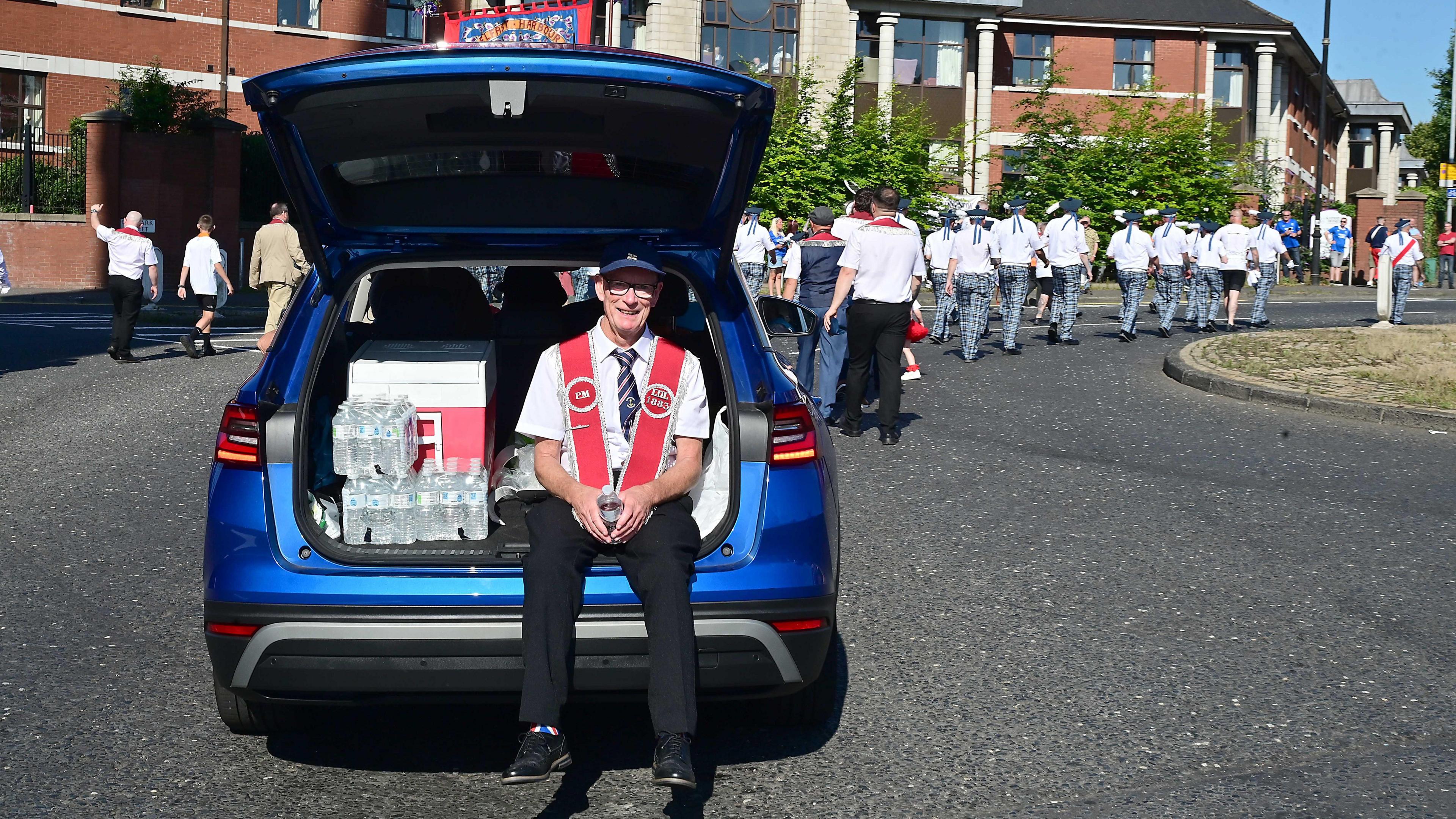 A man is sitting in the boot of a blue car next to a case of bottled water. He is wearing a dark blue cap and a red sash.