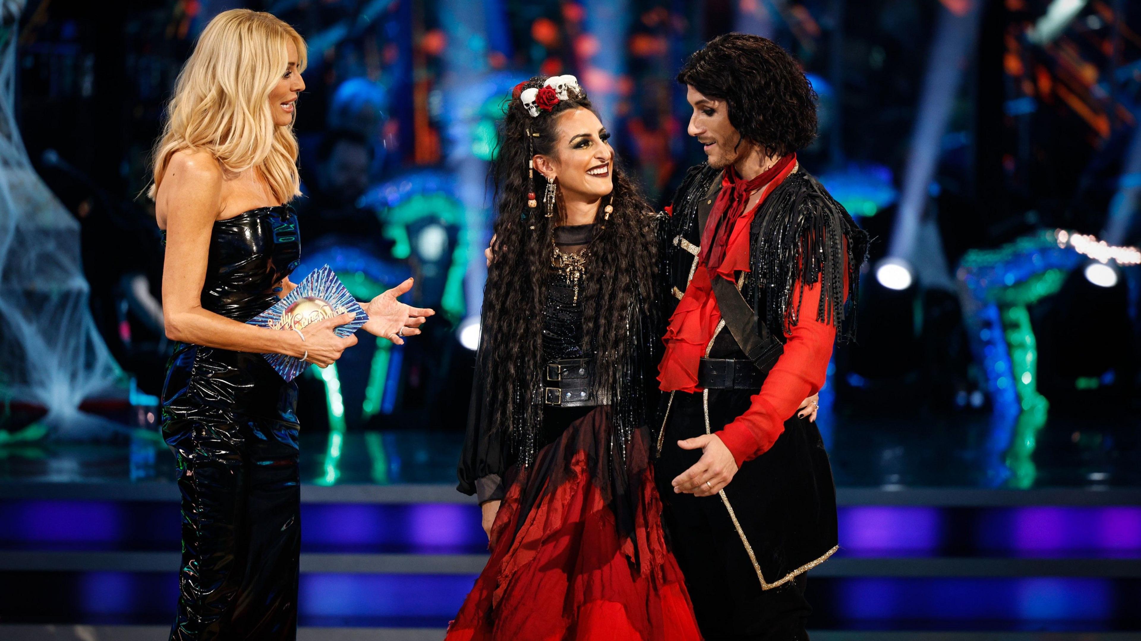 A woman in a black strapless dress smiling as she addresses a woman in a red and black dress and her partner. The woman in red has a skull and flower headpiece and a long black wig. Her partner is wearing a black jacket with gold trim and red accents. The couple is standing on a stage with a blurred background of lights and a crowd.