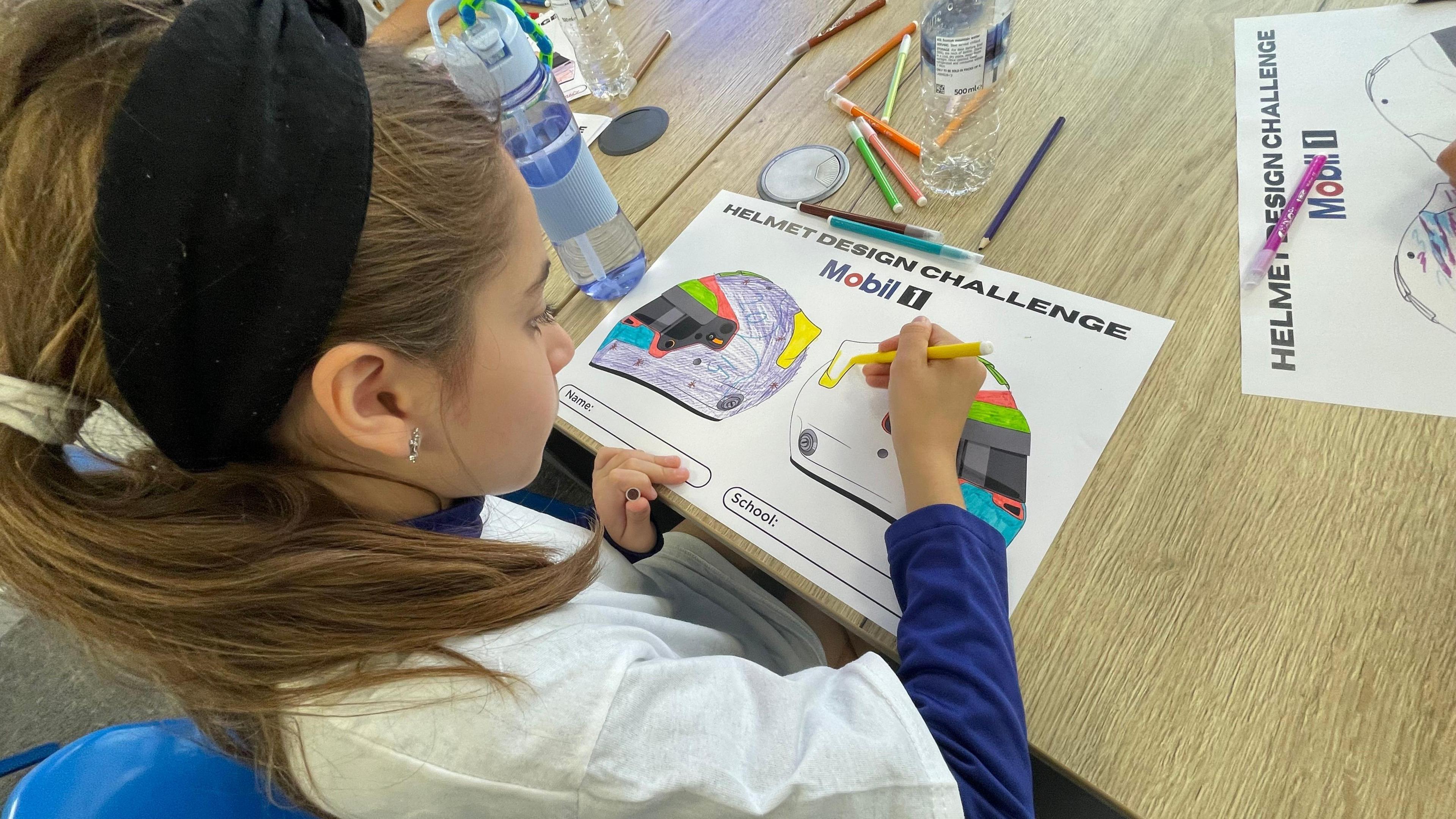 A girl with brown hair in a ponytail designing a motorsport helmet. The picture is taken from behind her shoulders. There are colour pens on the table and bottles of water.