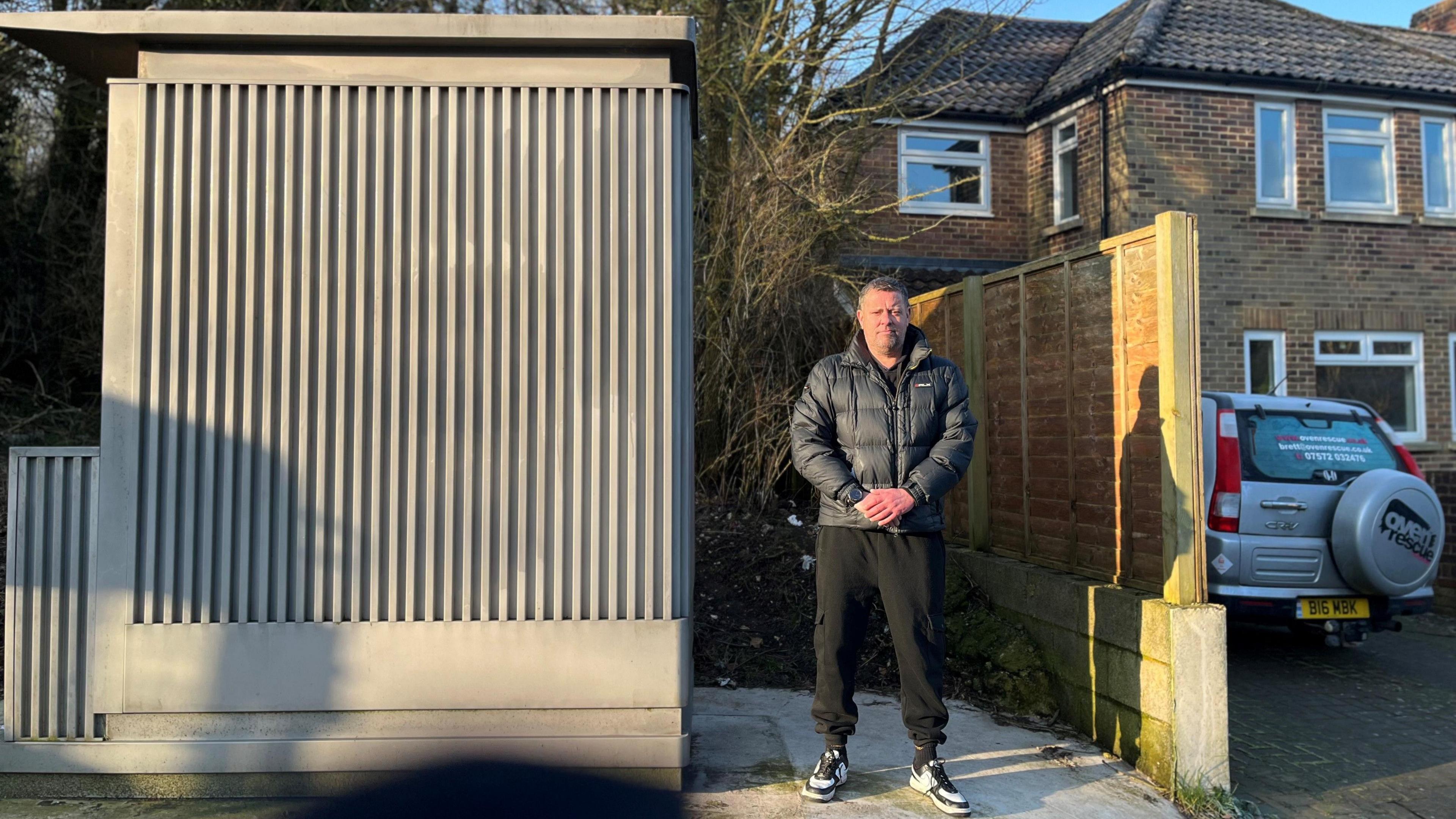 Brett Kemp, a man with short grey hair wearing a dark puffer jacket, standing next to the metal shed-style structure with corrugated sides. His house can be seen in the background