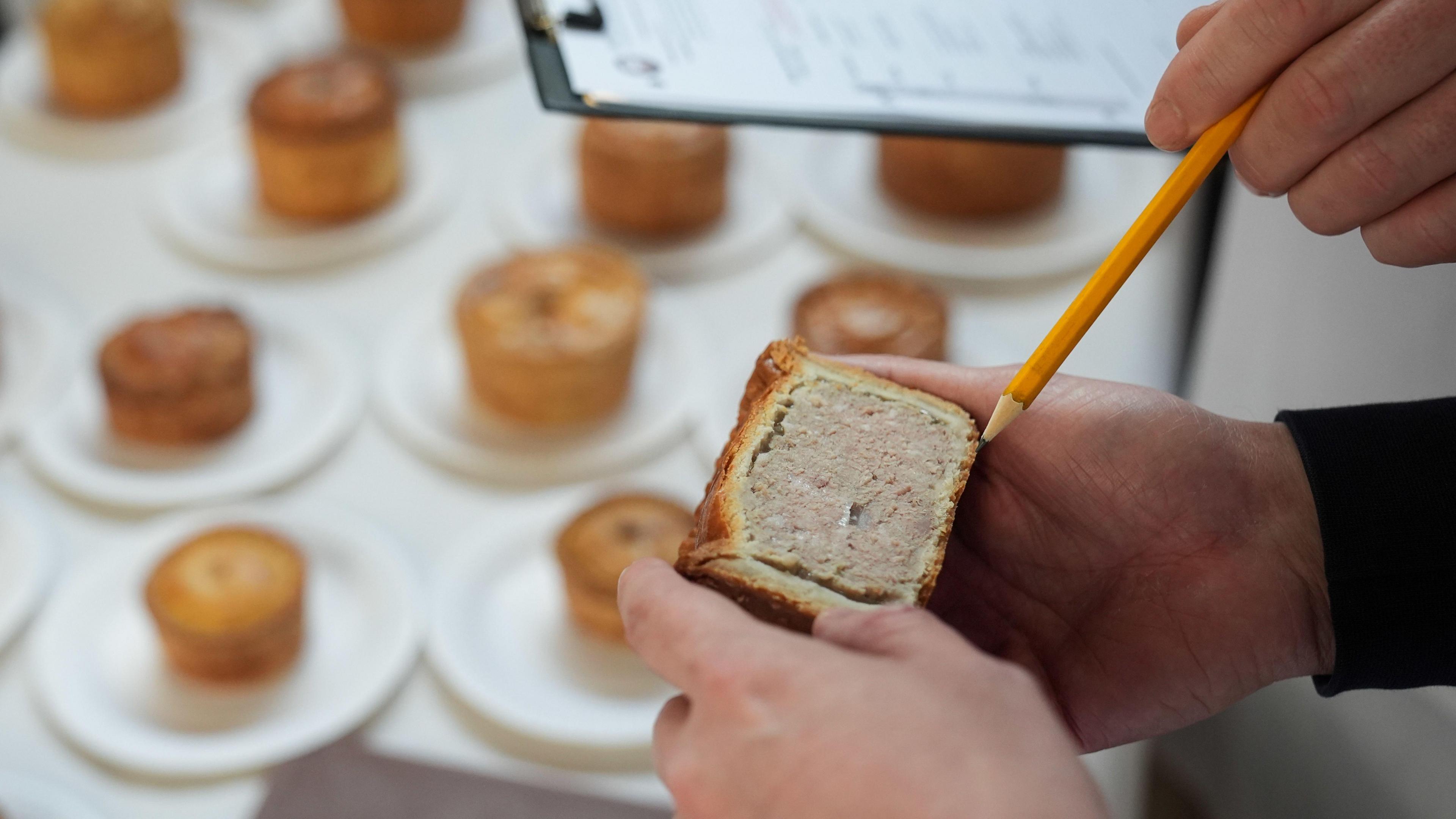 A photo of someone holding half a pork pie, and a pencil is pointing at it. In the background, underneath are more pies on plates on a table. 