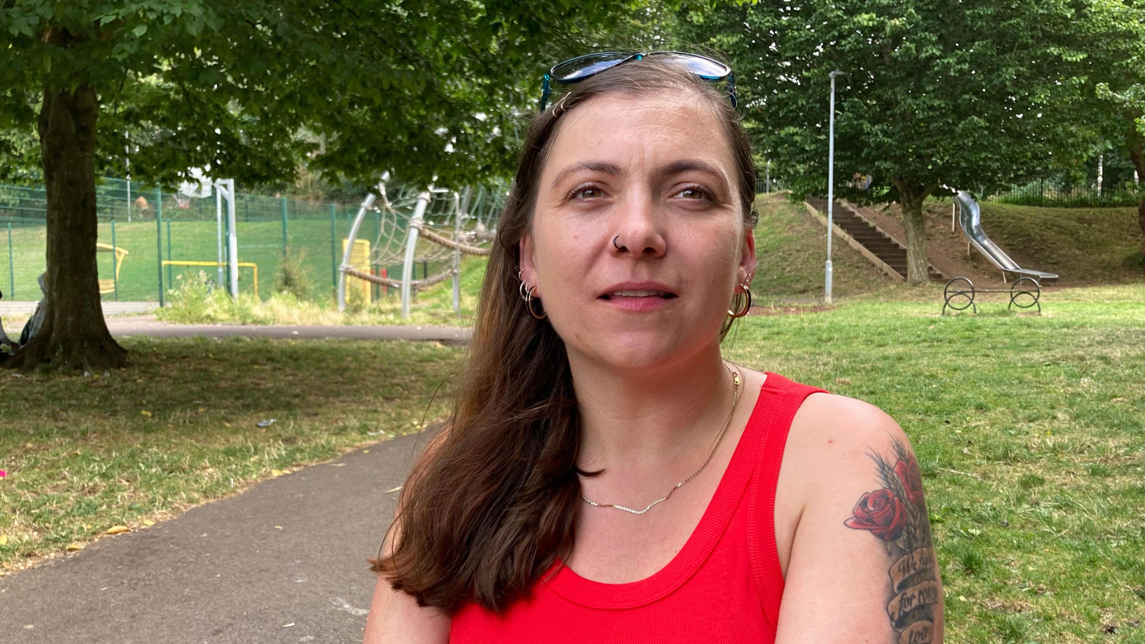 Toni Mayo pictured in a park, smiling at the camera. She has long dark hair and wears a red tank top and sunglasses on her head. She has a large tattoo with roses on her arm. 