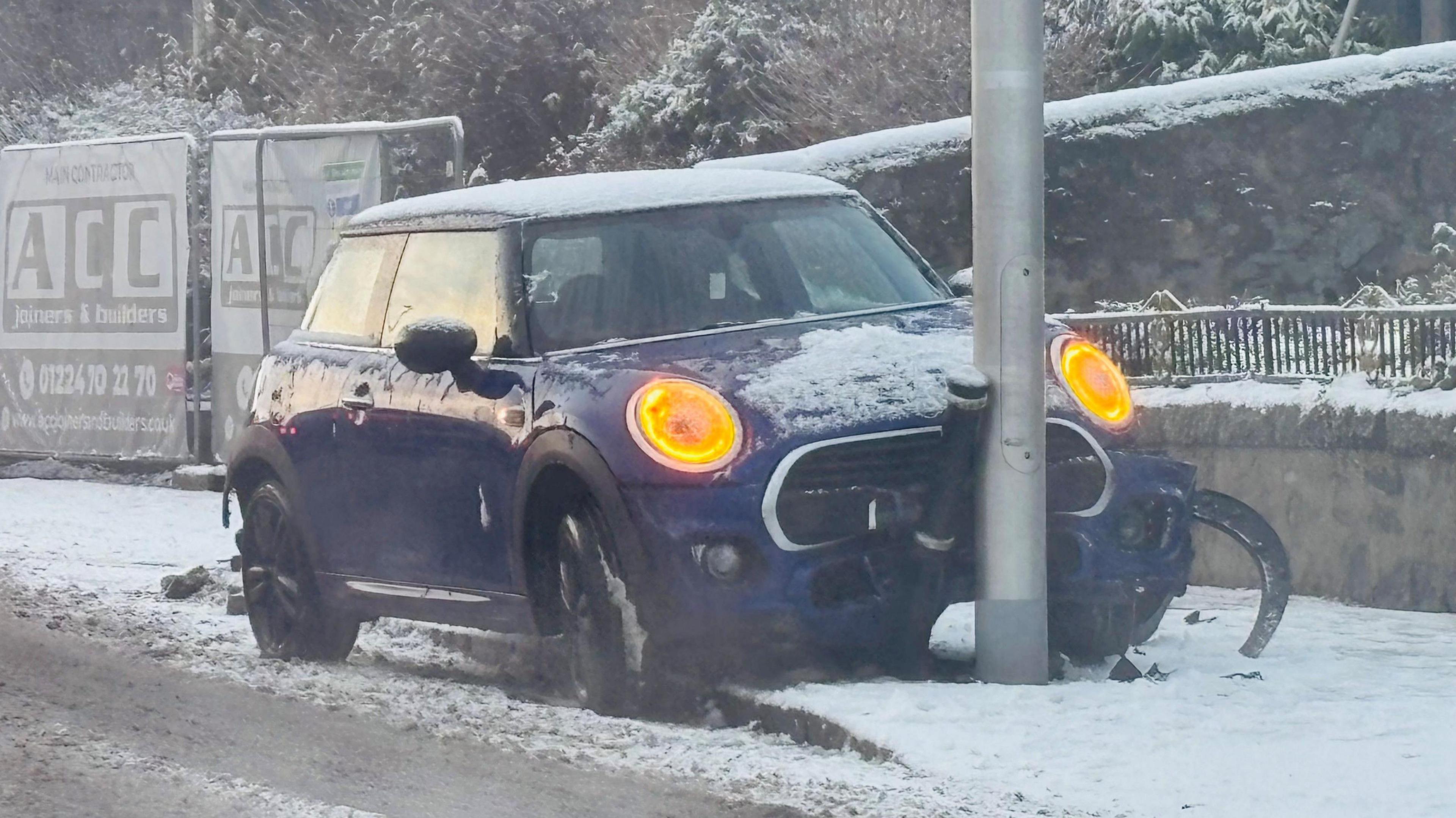 A blue mini crashed with orange lights on crashed into a bollard and lamppost in snow.