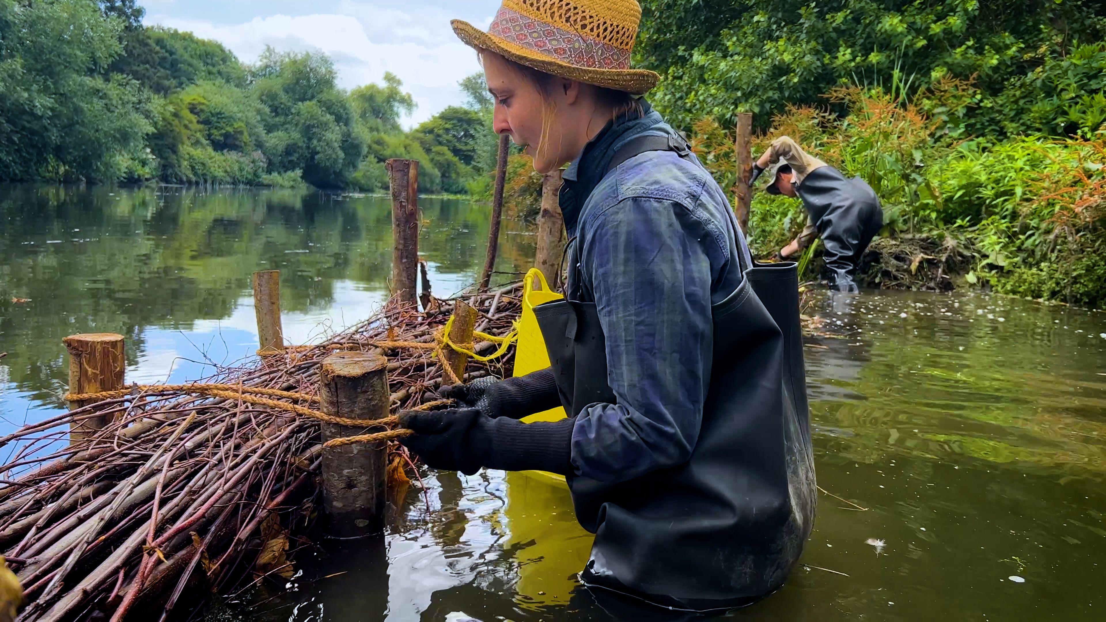 Two people in waterproof overalls build a wooden and branch barrier in a shallow river, surrounded by greenery. One ties rope around the structure, while the other secures materials in the background.