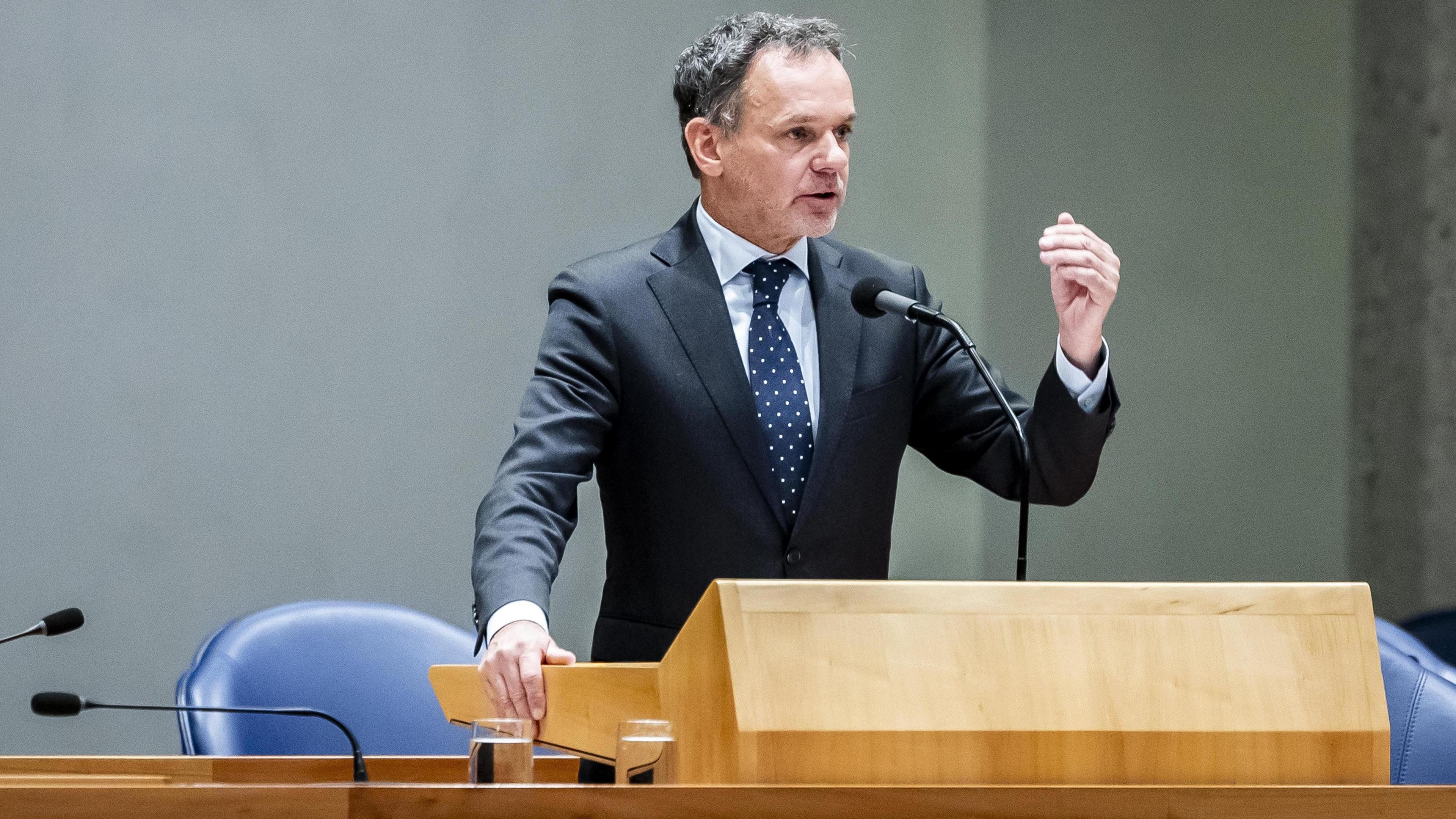 Dutch foreign minister Caspar Veldkamp gestures with his hand as he speaks at a wooden lectern in the Hague. He wears a grey suit, a white polka dot navy tie and white shirt and has short dark grey hair