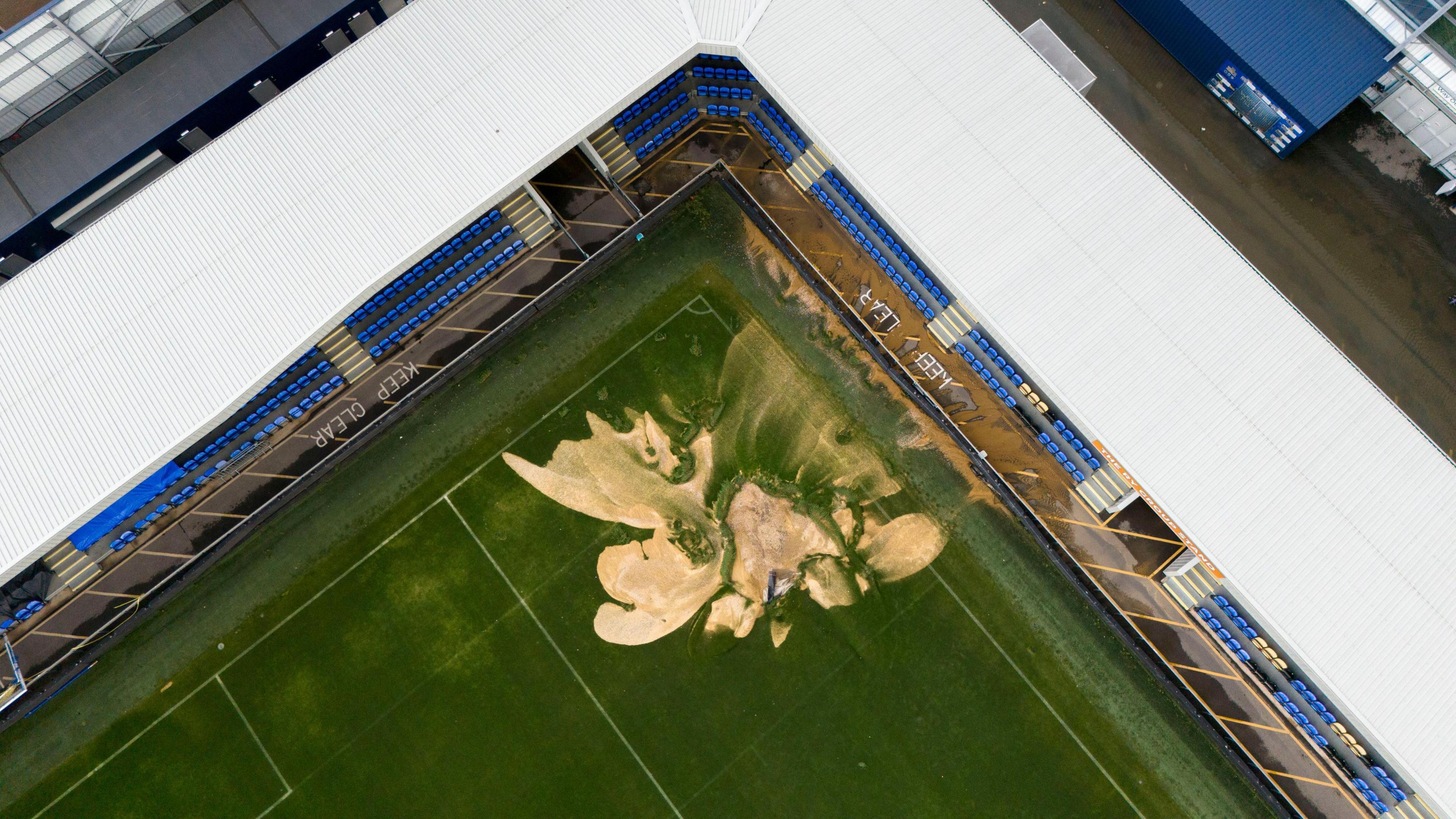 An aerial view of the football pitch at Wimbledon stadium in south-west london. In the middle, a sinkhole which looks like large sandy patch renders the pitch unusable. 