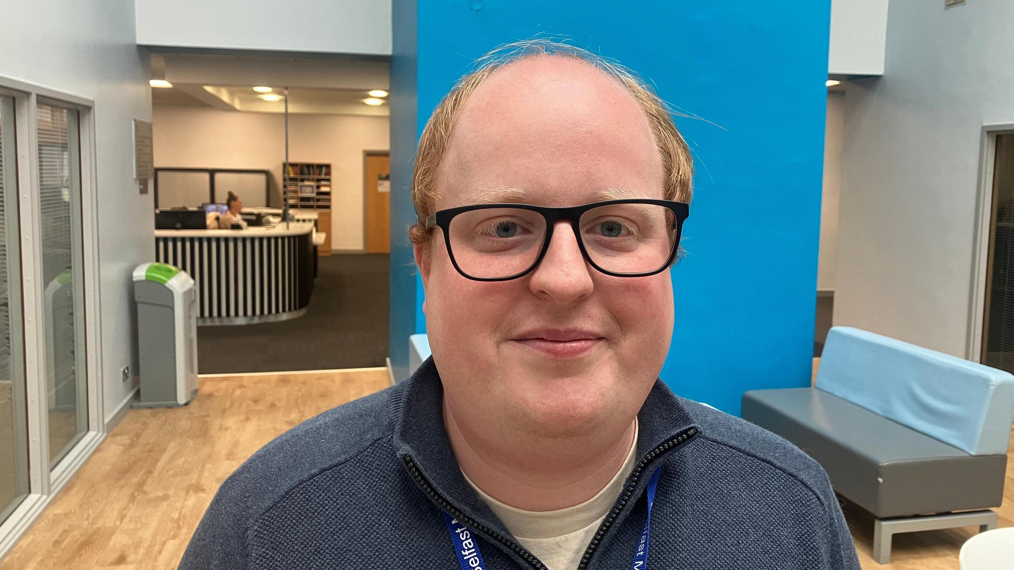 Michael is wearing glasses and a grey fleece. He is stood at a reception area in front of a blue wall and a grey sofa.