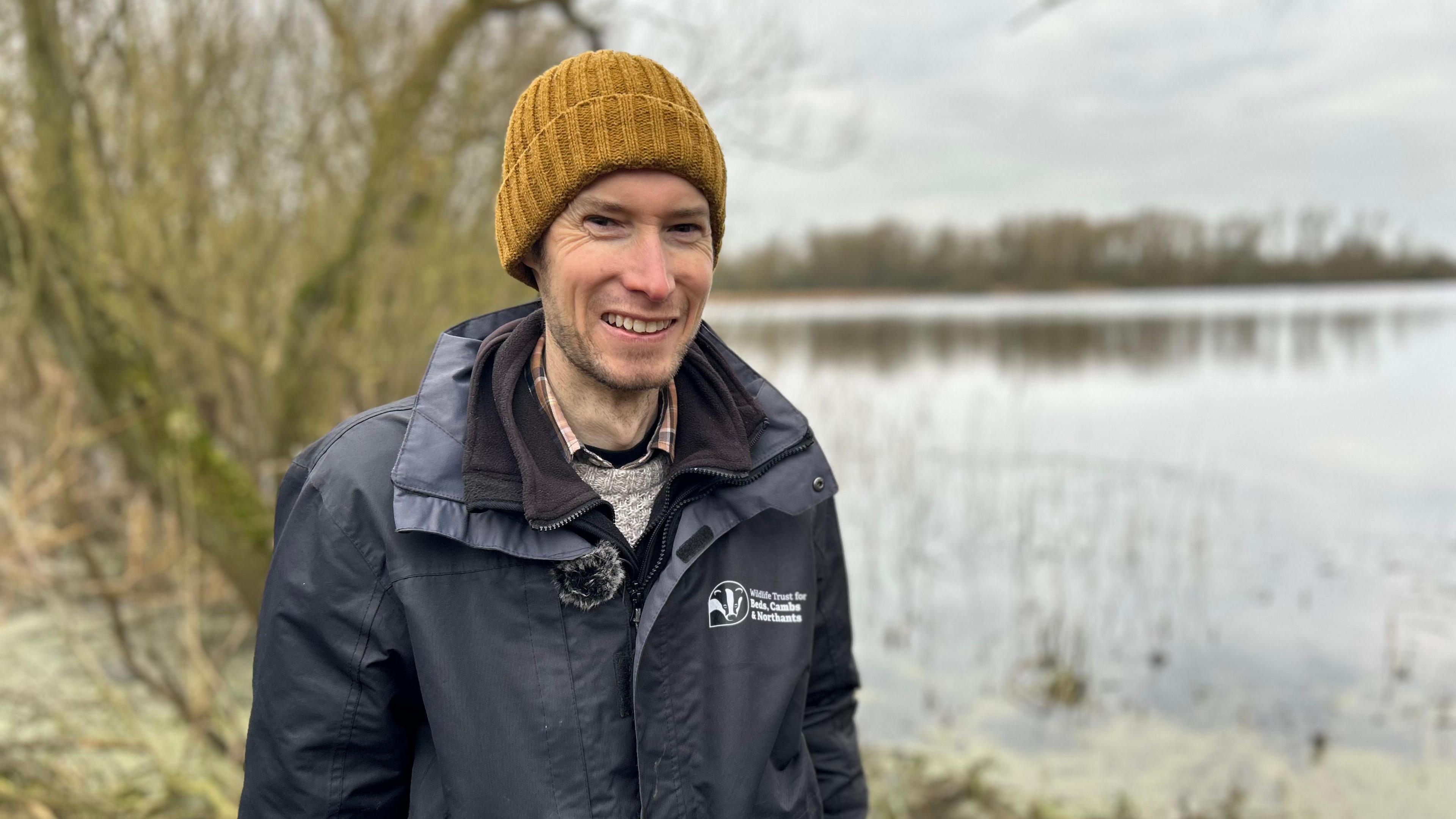 Matt Johnson in a navy raincoat wearing a yellow beanie standing beside Rushden Lakes.