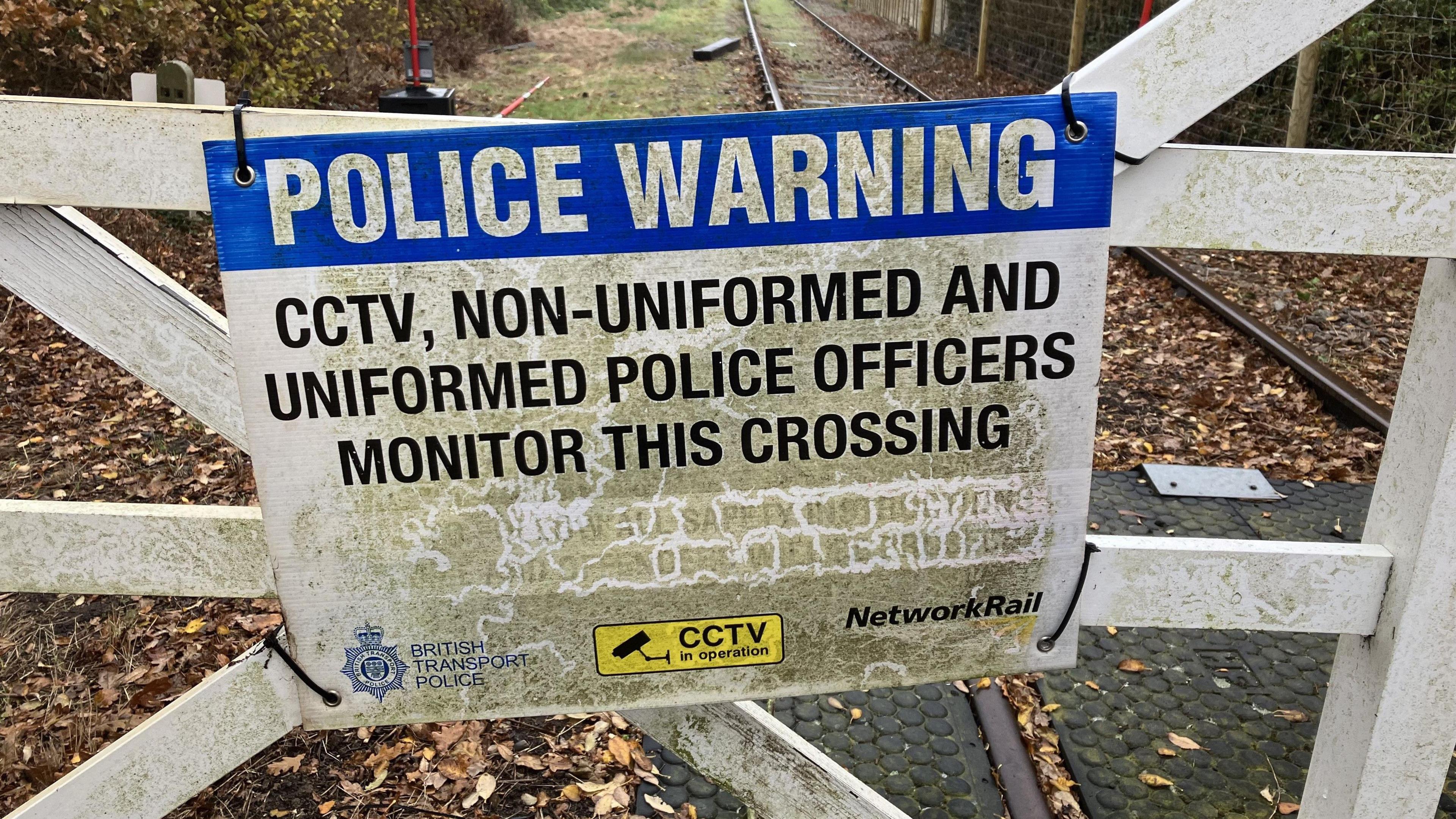 A blue and white sign reading "Police warning, CCTV, non-uniformed and uniformed police officers monitor this track" can be seen tied to crossing gates near railway line   