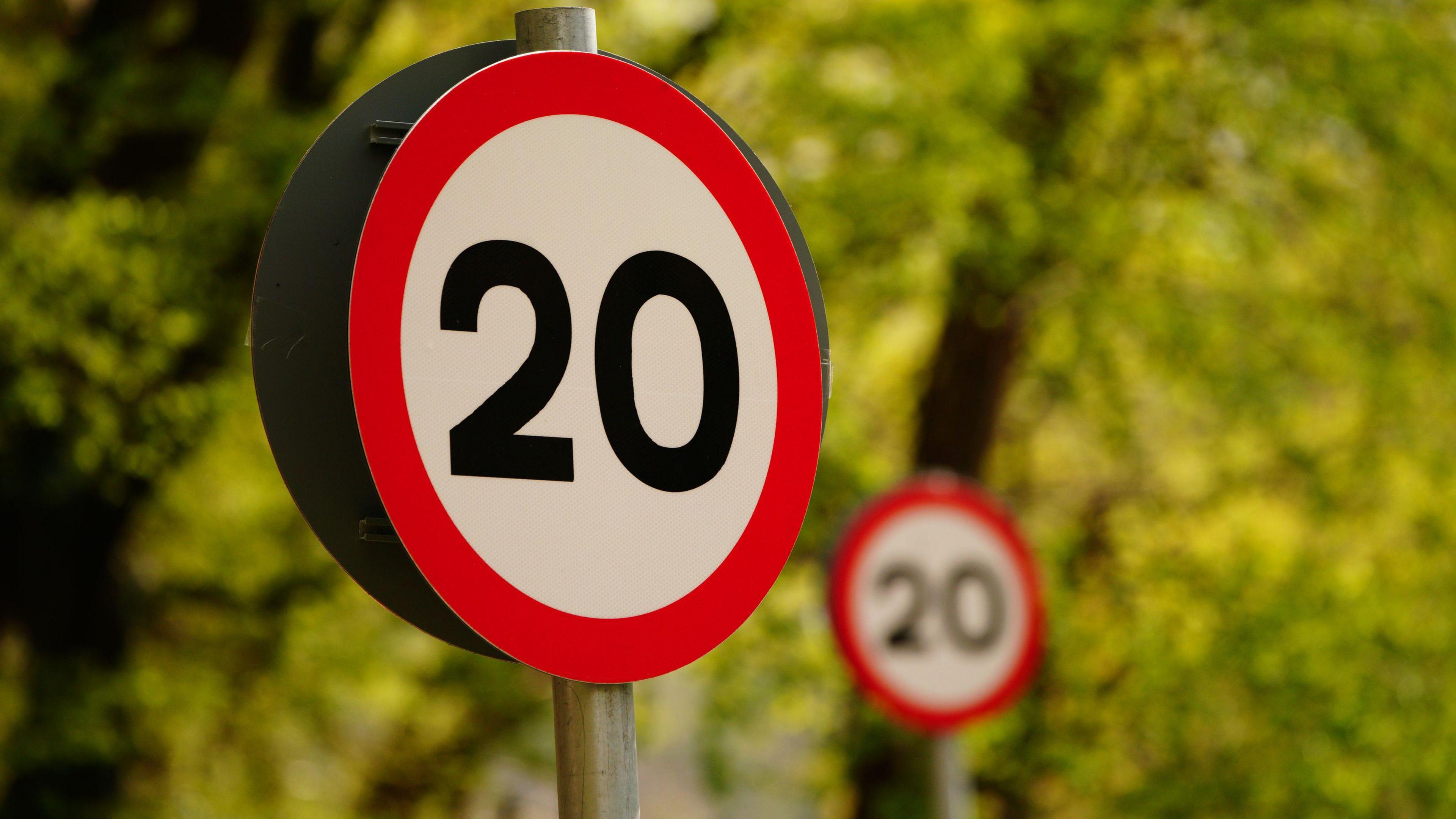 A round road sign with the 20 in black on a white background with a red circle around the edge. The sign is in front of another identical one, which is blurred in the background in front of lots of trees.