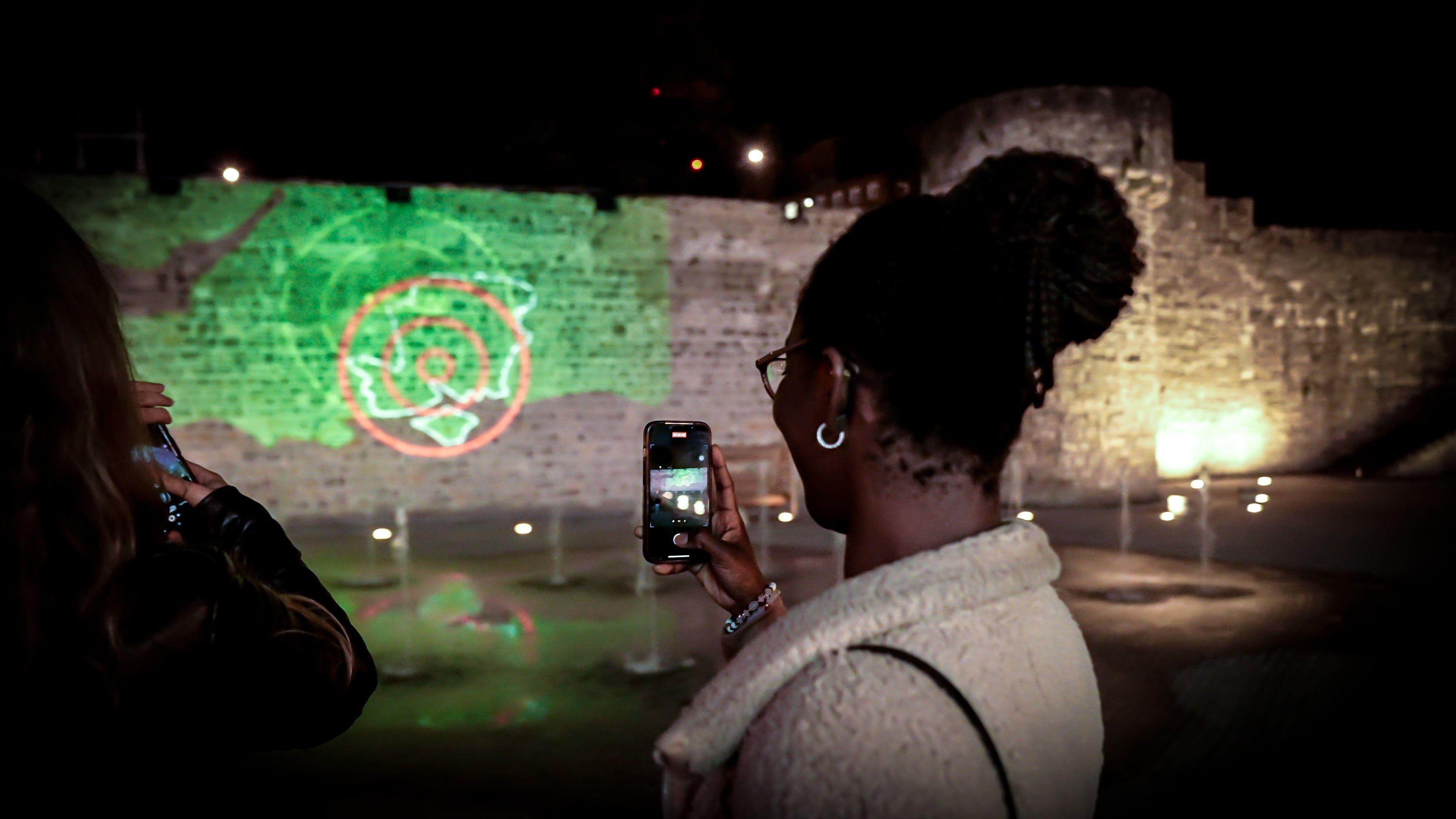 A woman with her black hair tied up in a bun is using her phone to take a picture of a green projection onto an old wall.