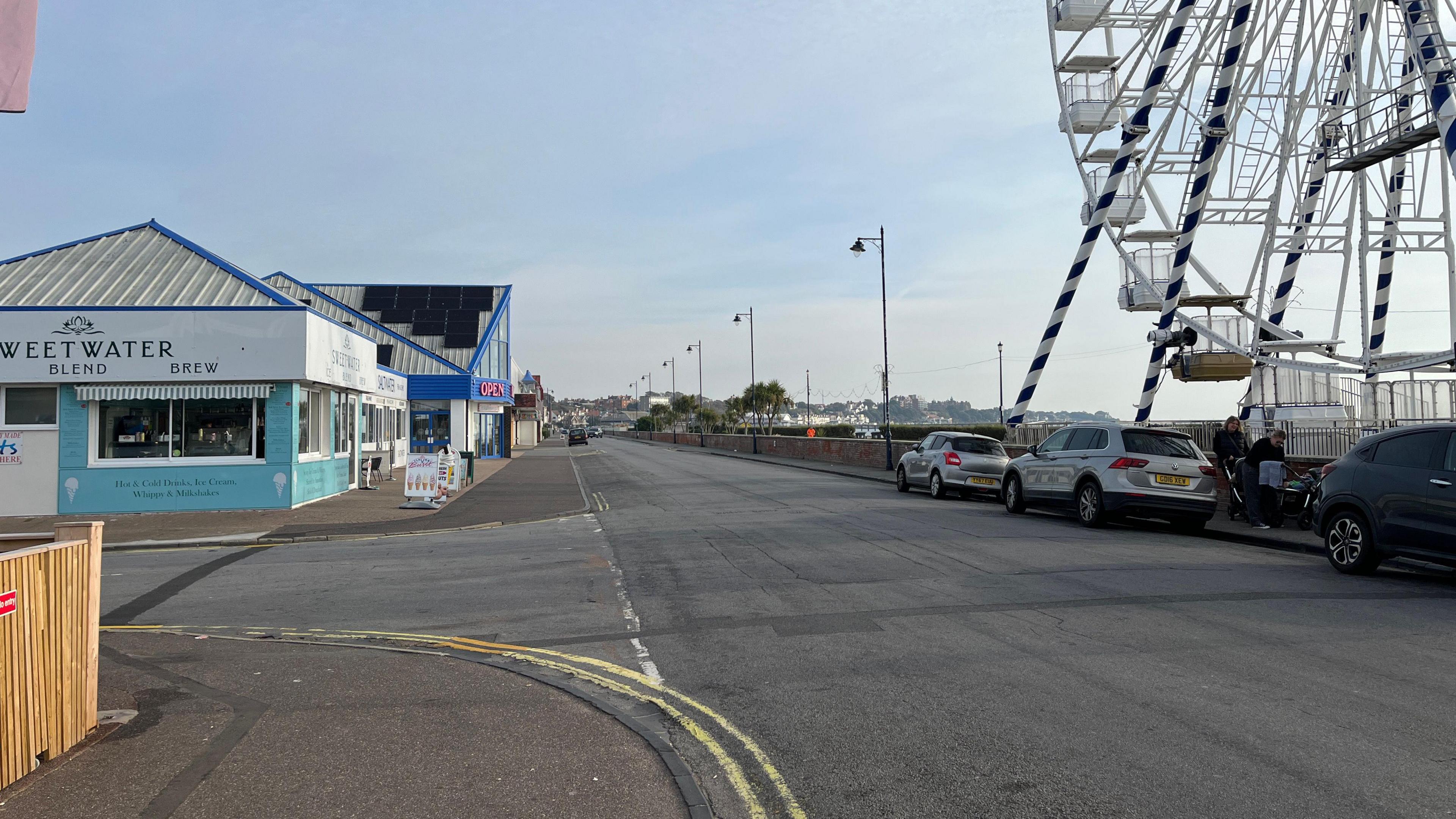 Sea Road in Felixstowe. Cars are parked on the right hand side of the road in front of a ferris wheel. Businesses line the opposite site of the road. 
