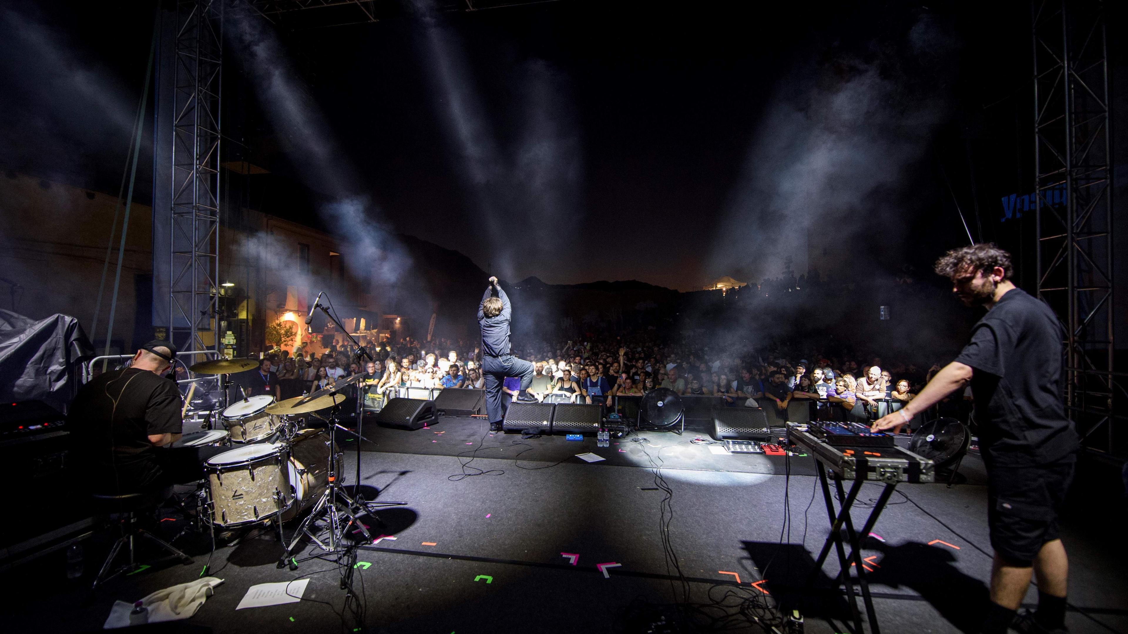 A band performing on stage. The photo is taken from behind the band, and in the distance you can see the audience. On the right, there is a man standing by a keyboard. On the left, there is a man sitting at a drum-set. In the middle, and further away, is a man standing, holding a microphone. All three are dressed all in black. 