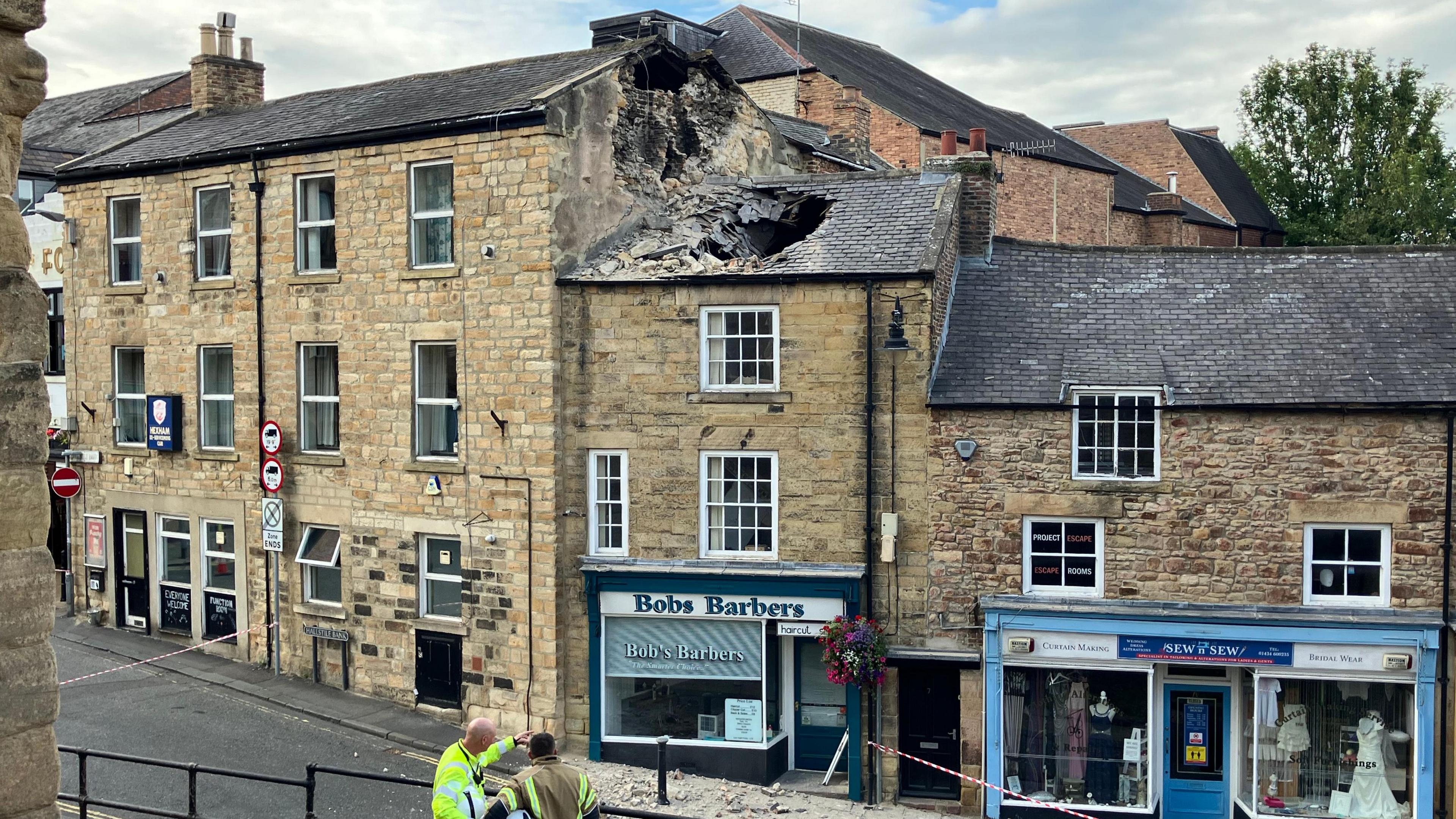 A view of a street of tall, terraced, sandstone buildings. The two to the right are shops, with one of them, Bobs Barbers has a gaping hole in the tiled roof due to a collapsed chimney. Some rubble is strewn on the ground in front of the building and the street is taped off. 
