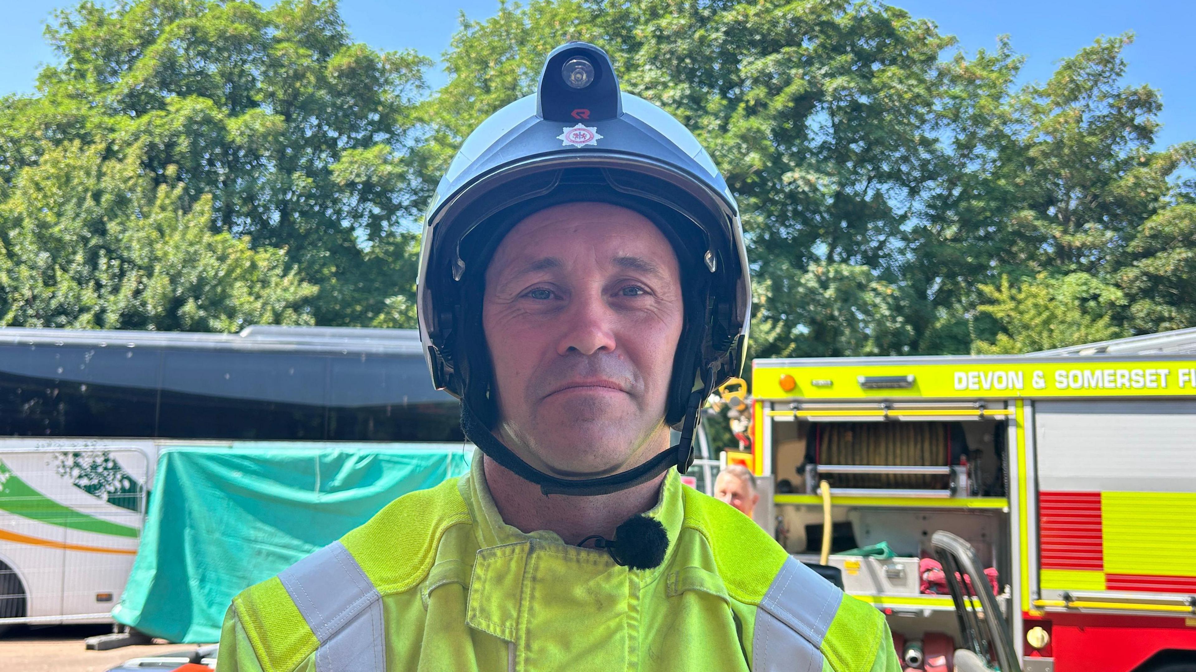 A firefighter with a yellow jacket and a dark coloured helmet. He has light stubble